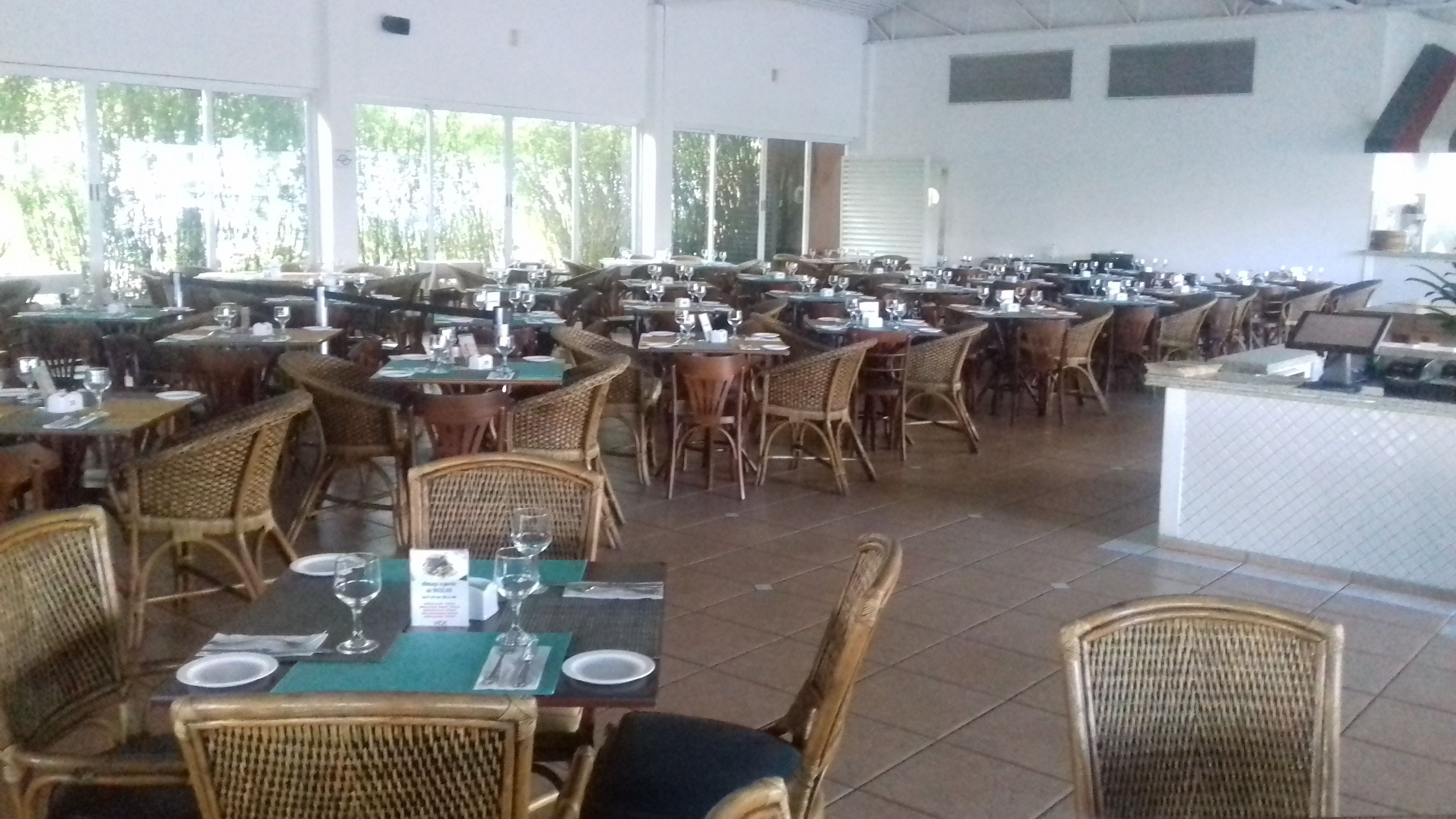 Een ruime restaurantruimte met meerdere tafels en rieten stoelen, grote ramen en een betegelde vloer.