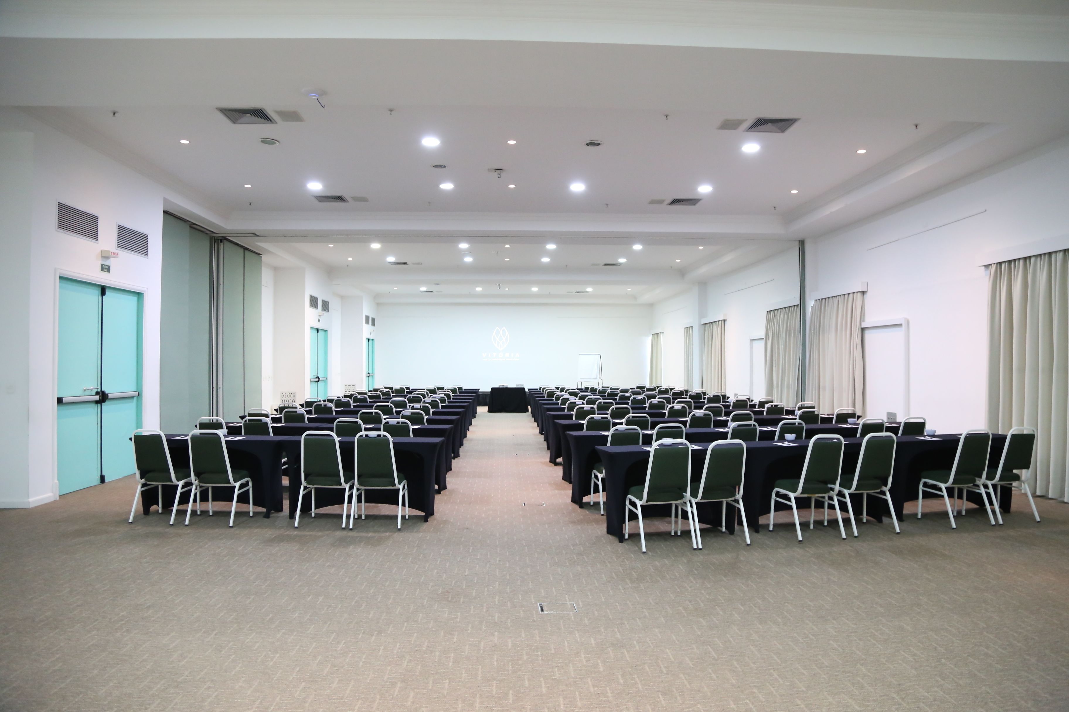 Une salle de conférence avec des rangées de tables et de chaises, un sol recouvert de moquette et un plafond blanc doté d’un éclairage encastré.