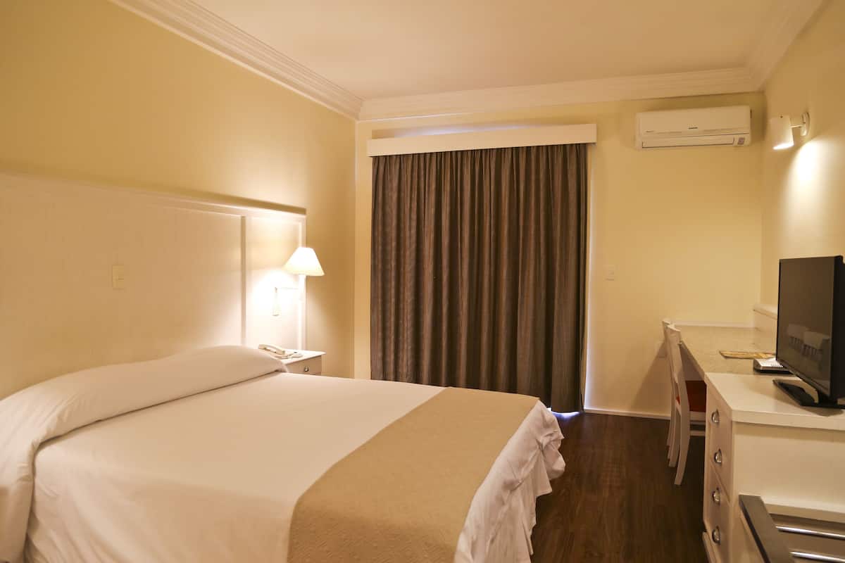 Een hotelkamer met een bed, een bureau met een televisie, een lamp en een airconditioning.