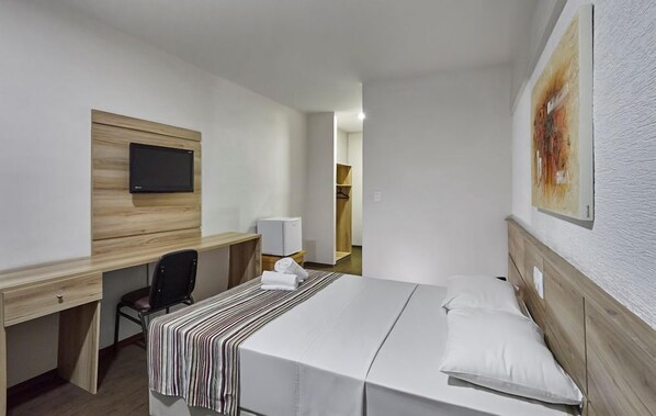A hotel room with a bed, a desk, a chair, a TV mounted on a wooden panel, and a painting on the wall.