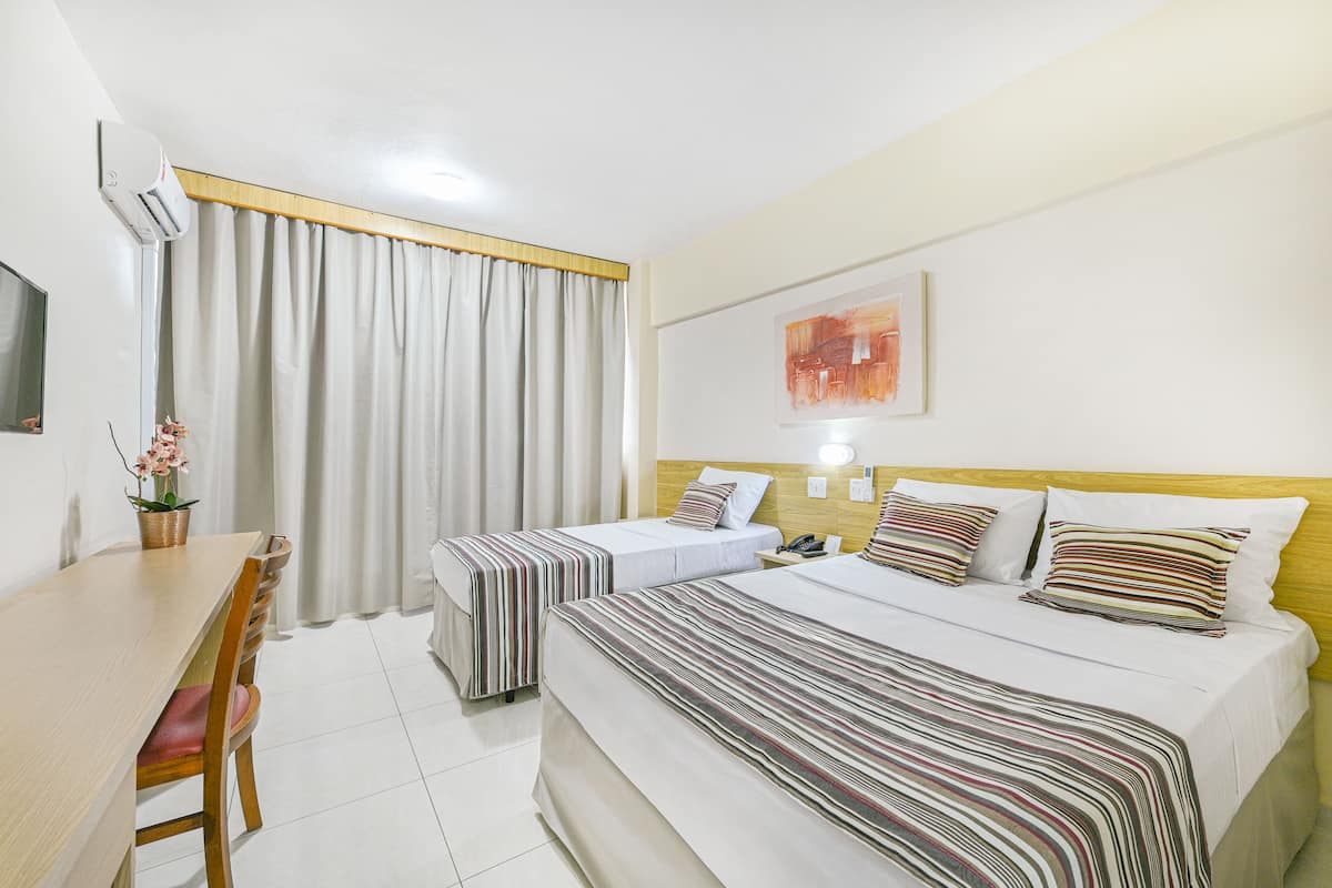 A hotel room with two beds, a wooden headboard, a dining table with chairs, and a window with curtains.