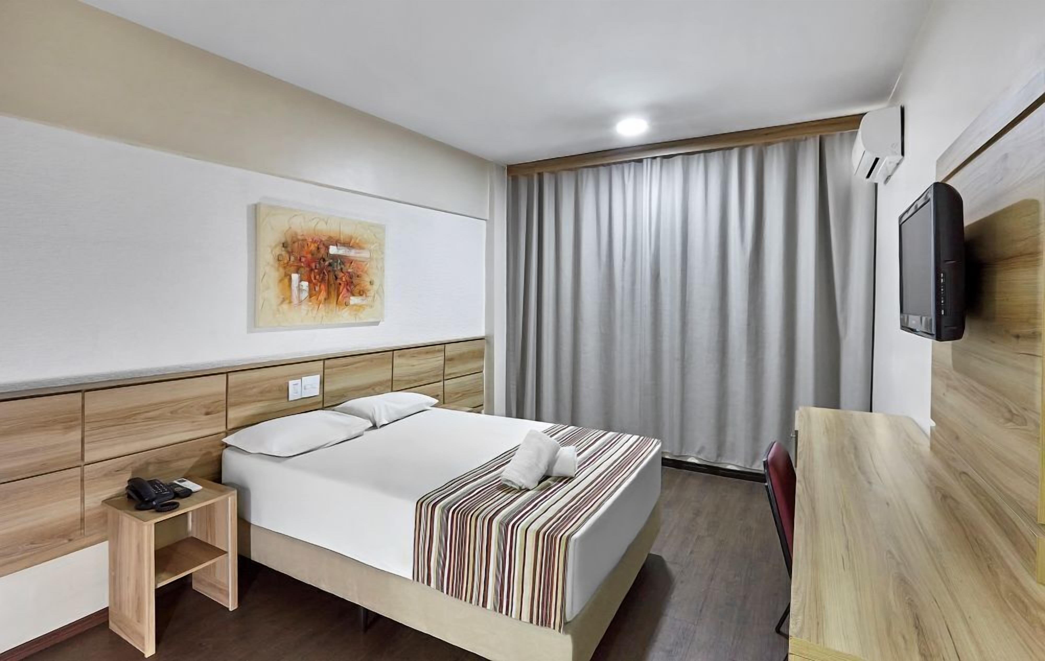 A hotel room with a bed, a bedside table with a phone, a TV mounted on the wall, and a large window with curtains.
