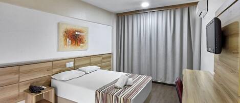 A hotel room with a bed, a bedside table with a phone, a TV mounted on the wall, and a large window with curtains.