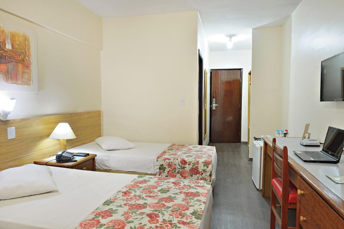 A hotel room with two beds, a desk with a laptop, a TV, and a door leading to another room.