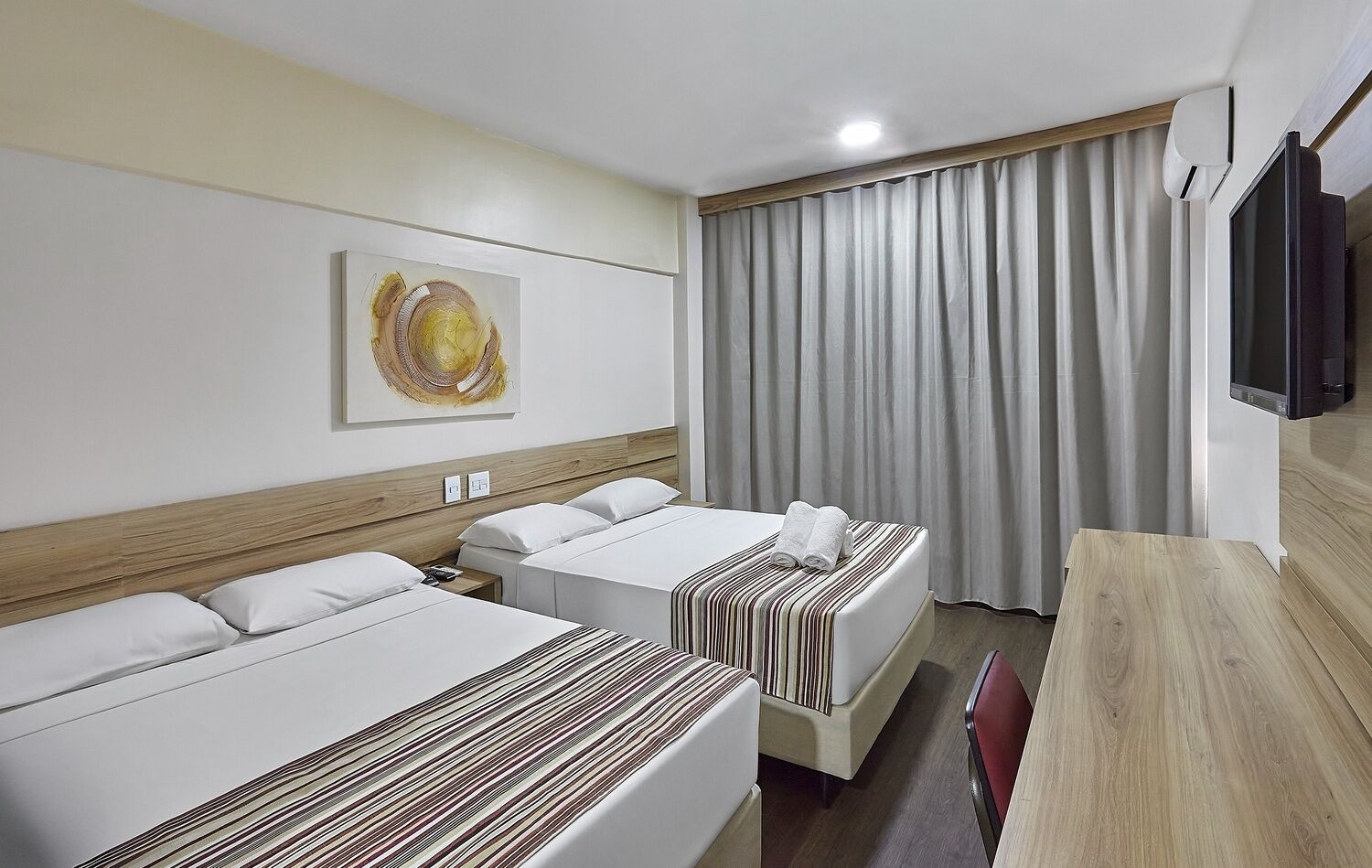 A hotel room with two beds, a wooden headboard, a flat-screen TV, a painting on the wall, and a large window with curtains.
