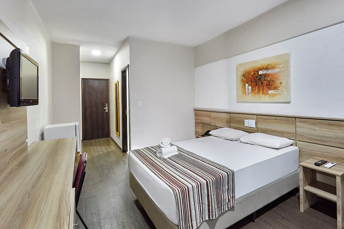 A hotel room with a bed, a bedside table with a phone, a TV mounted on the wall, and a painting above the bed.