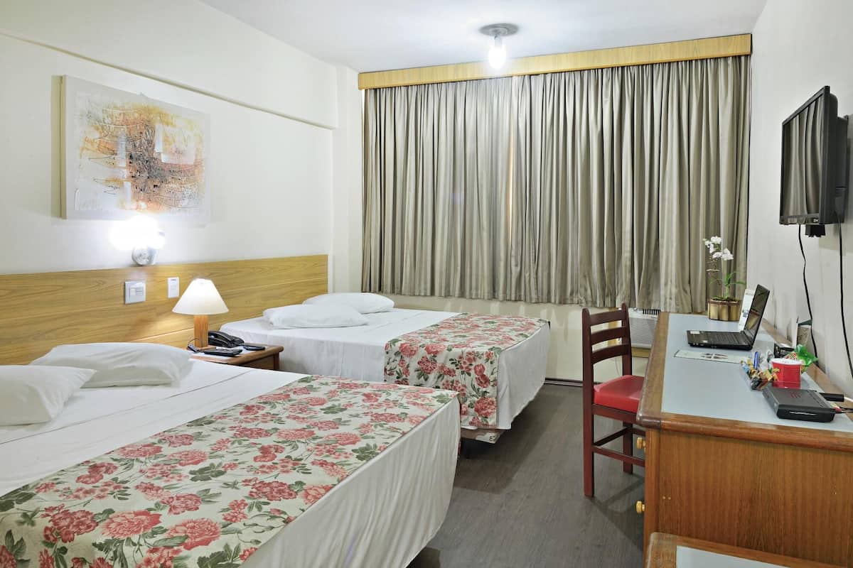 A hotel room with two beds, a desk with a laptop, a chair, a TV, and a window with curtains.