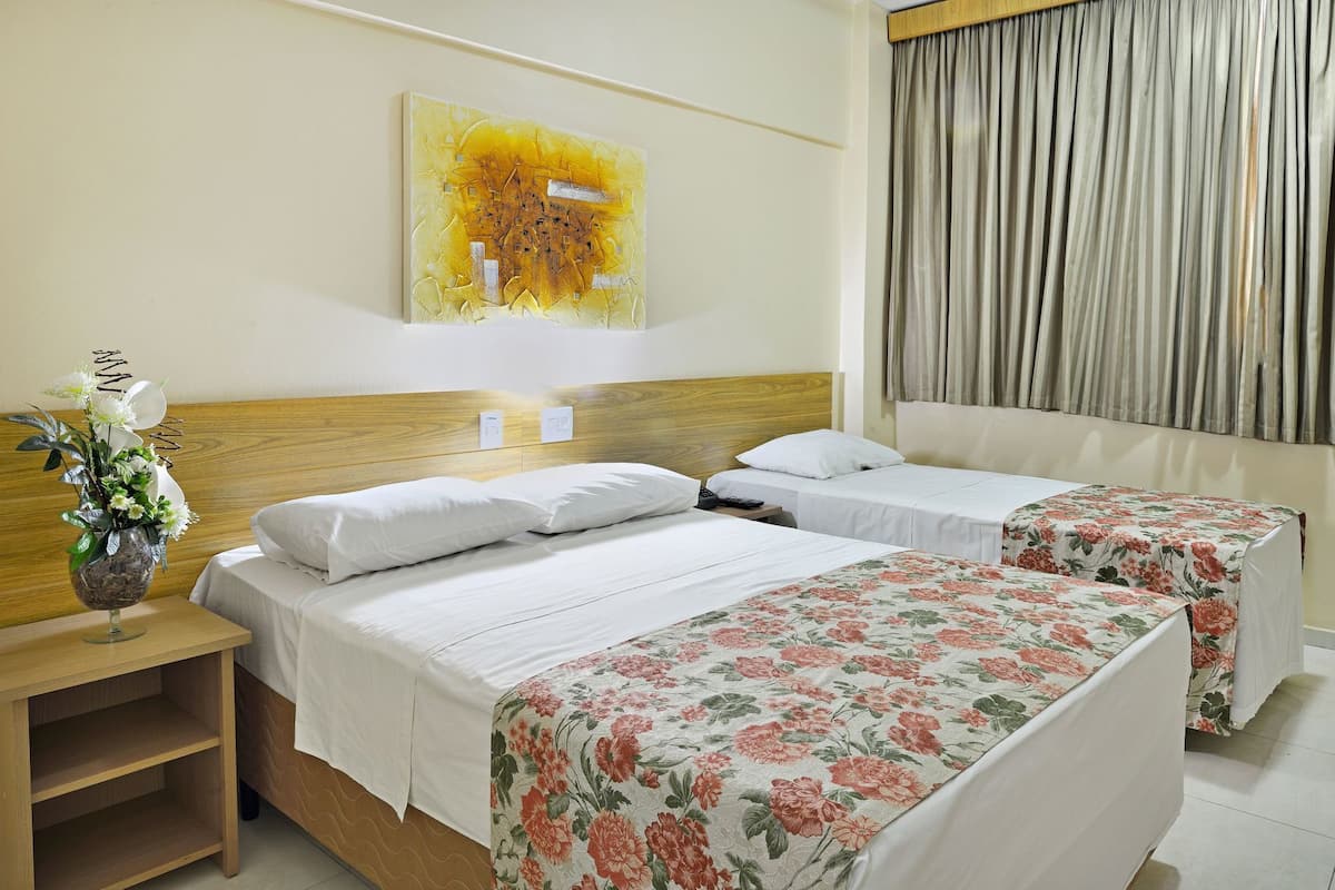 A hotel room with two beds, a wooden headboard, a floral bedspread, a painting on the wall, a nightstand with a vase of flowers, and a window with curtains.