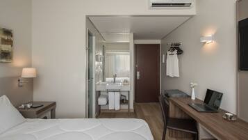 Quarto de hotel moderno com cama, escrivaninha, laptop, cadeira, mesa de cabeceira, luminária, espelho, guarda-roupa e banheiro com pia e chuveiro.