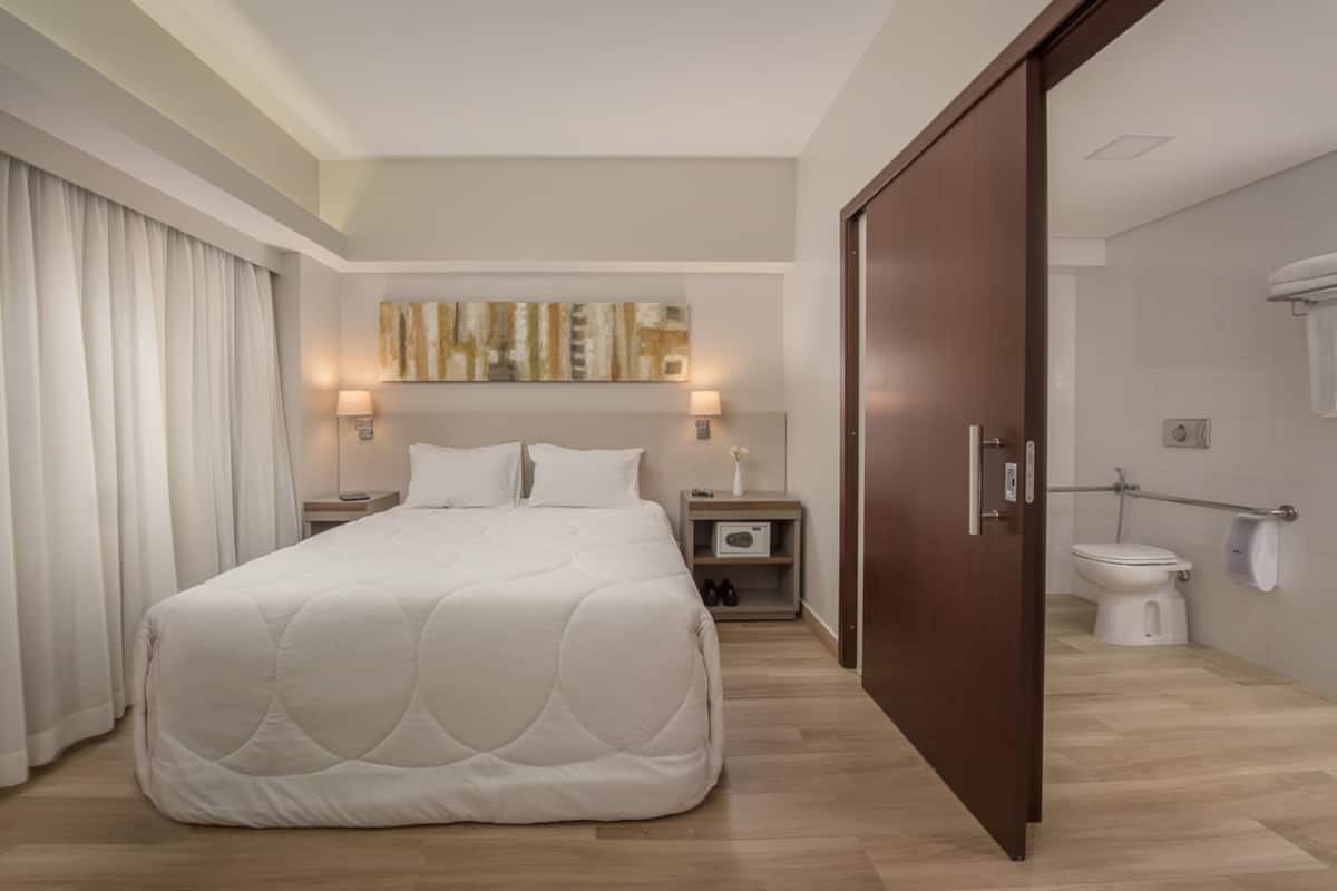 A hotel room with a large bed, a nightstand, a wall-mounted lamp, a painting, a wooden floor, and a bathroom with a toilet and shower.