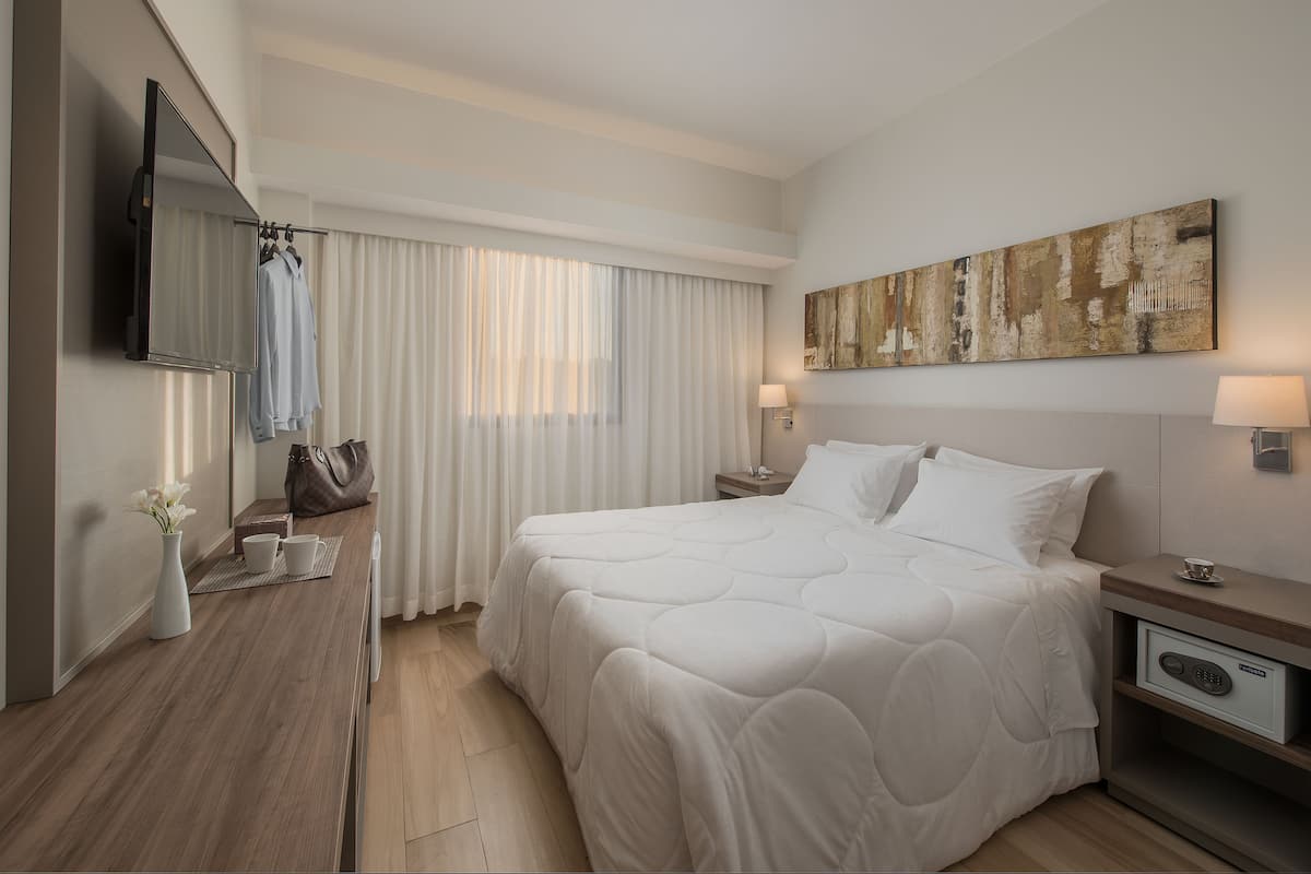 A modern hotel room with a large bed, a flat-screen TV, a bedside table with a lamp, and a painting on the wall.