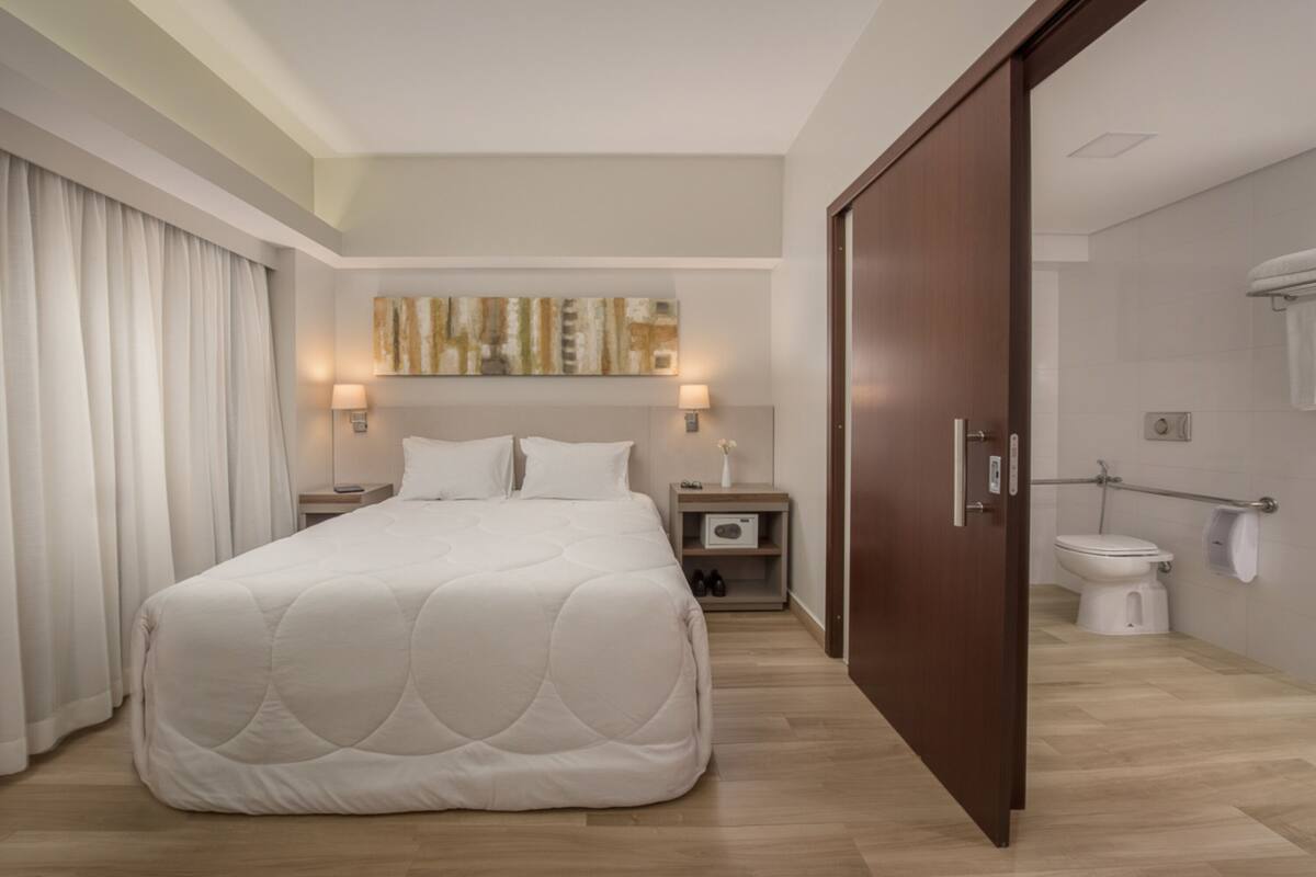 A hotel room with a large bed, a nightstand, a wall-mounted lamp, a painting, a door, and a bathroom with a toilet and shower.