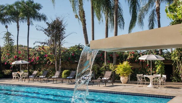 A swimming pool with a water feature, surrounded by lounge chairs and umbrellas, with palm trees and flowering plants in the background. - Hotel Nacional Distributed by Intercity (São José do Rio Preto)