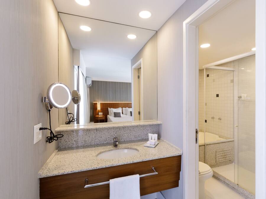 Une salle de bain moderne avec un lavabo sur un plan de travail en granit, un meuble-lavabo en bois, un grand miroir rond avec éclairage intégré, une cabine de douche en verre et, au fond, une chambre à coucher visible.