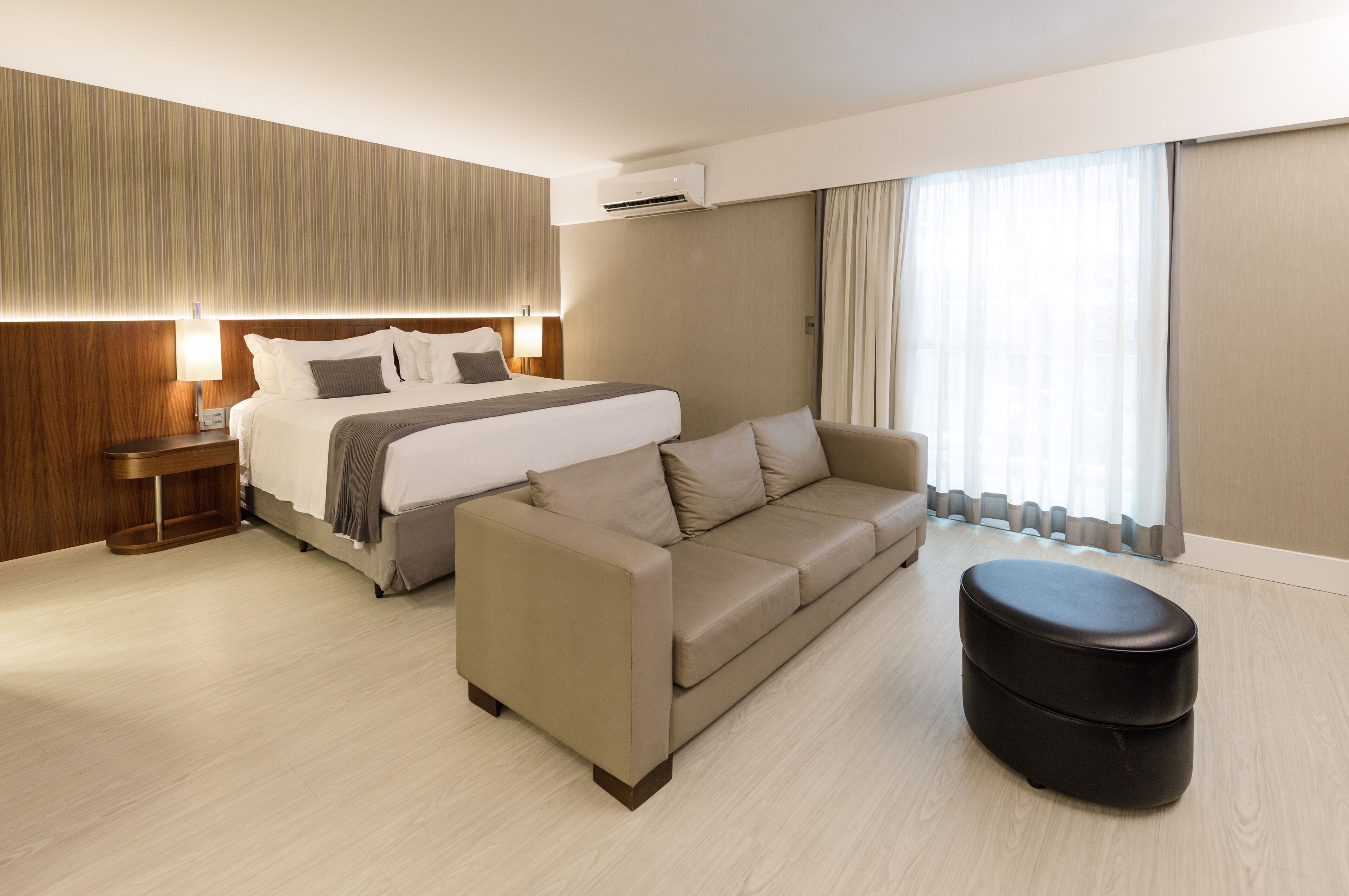 Quarto de hotel moderno com uma cama grande, um sofá, um pequeno banquinho e uma mesinha de cabeceira.