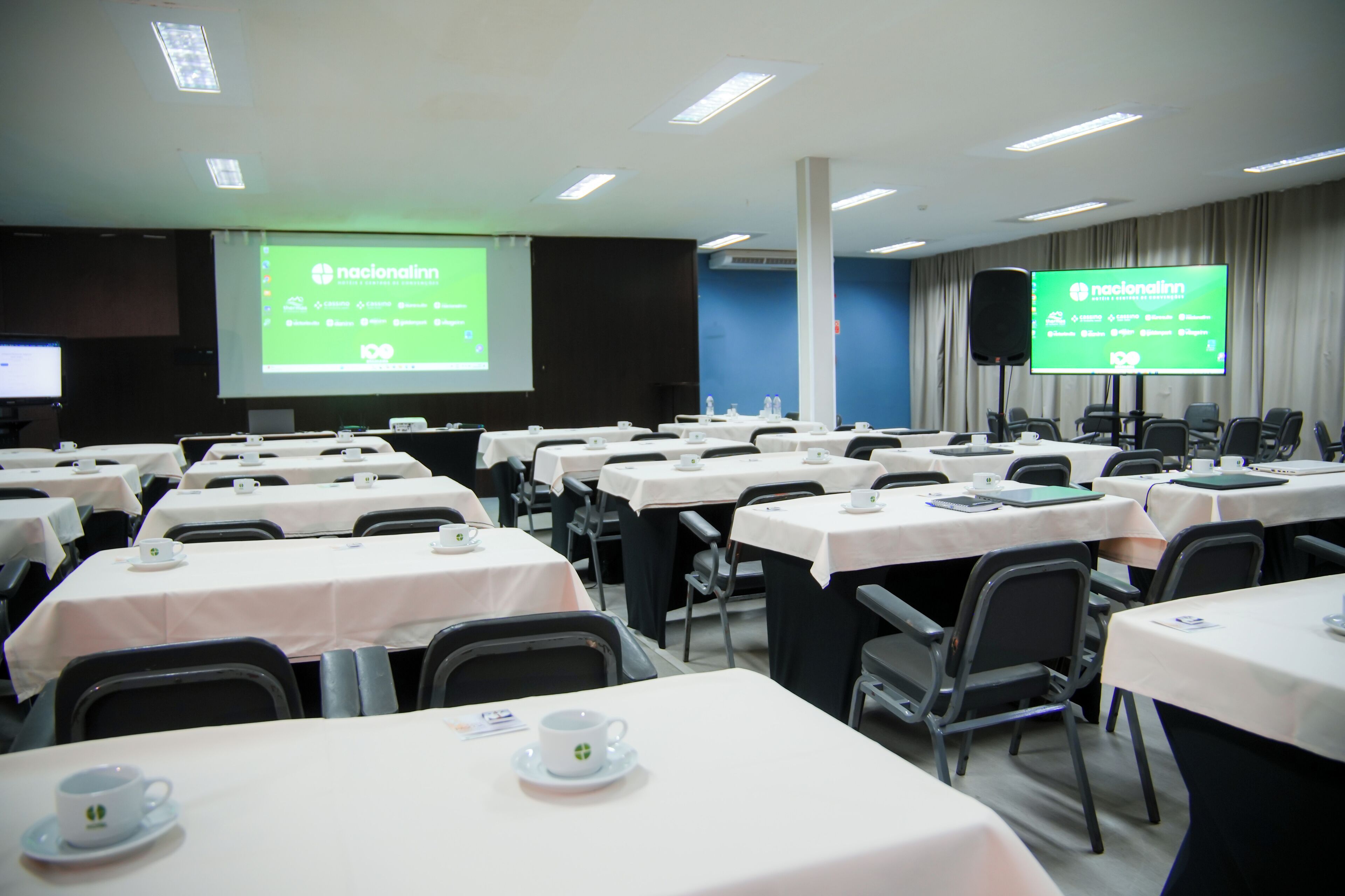 Un salón de conferencias con varias mesas, cada una con mantel blanco y una taza. Dos pantallas grandes muestran el mismo contenido.