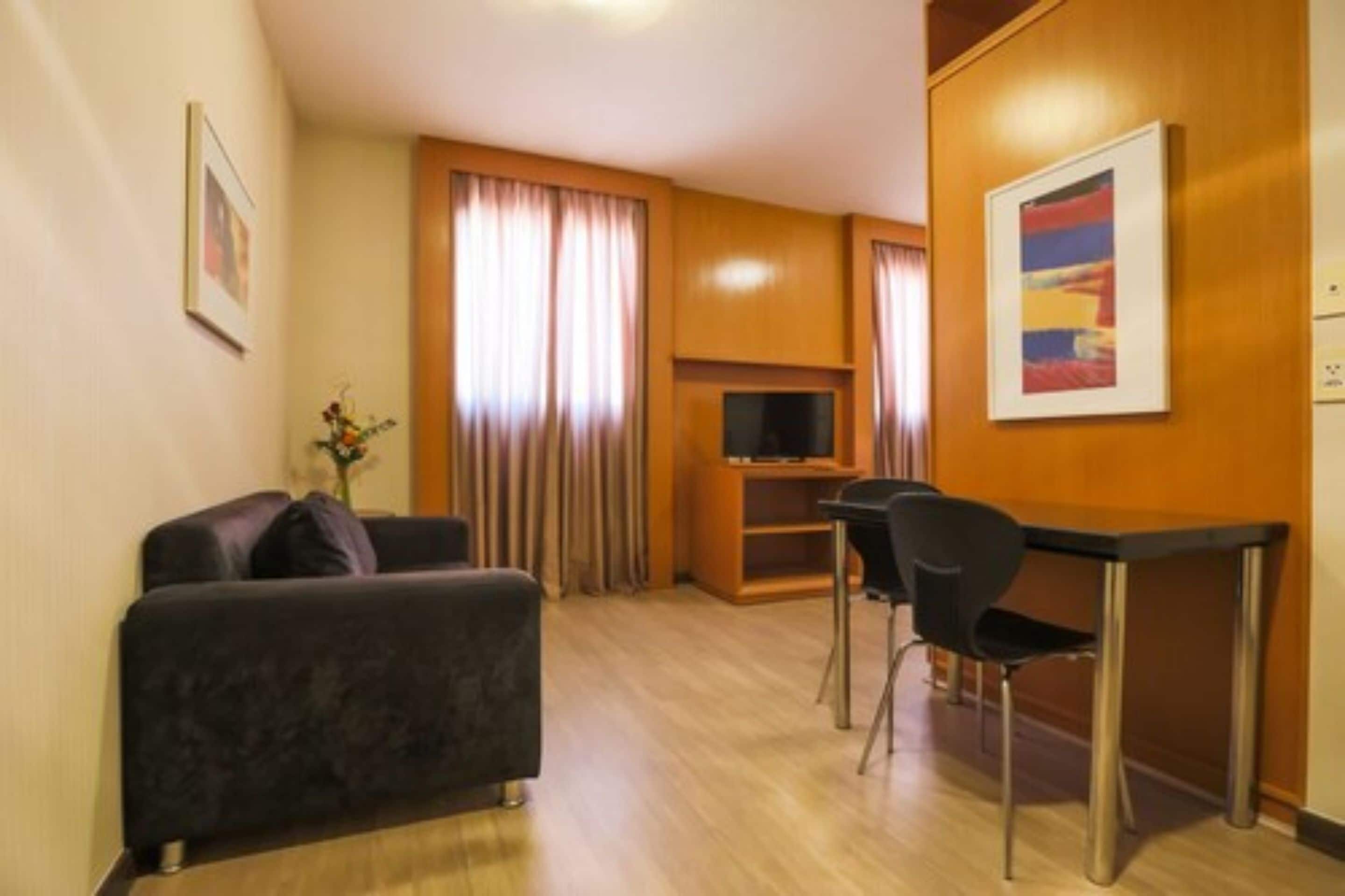 Quarto de hotel compacto com sofá, uma televisão pequena, secretária com duas cadeiras e um armário de madeira.
