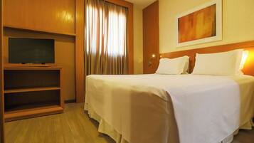 Habitación de hotel con una cama, televisión en un mueble bajo, un cuadro en la pared y una ventana con cortinas.