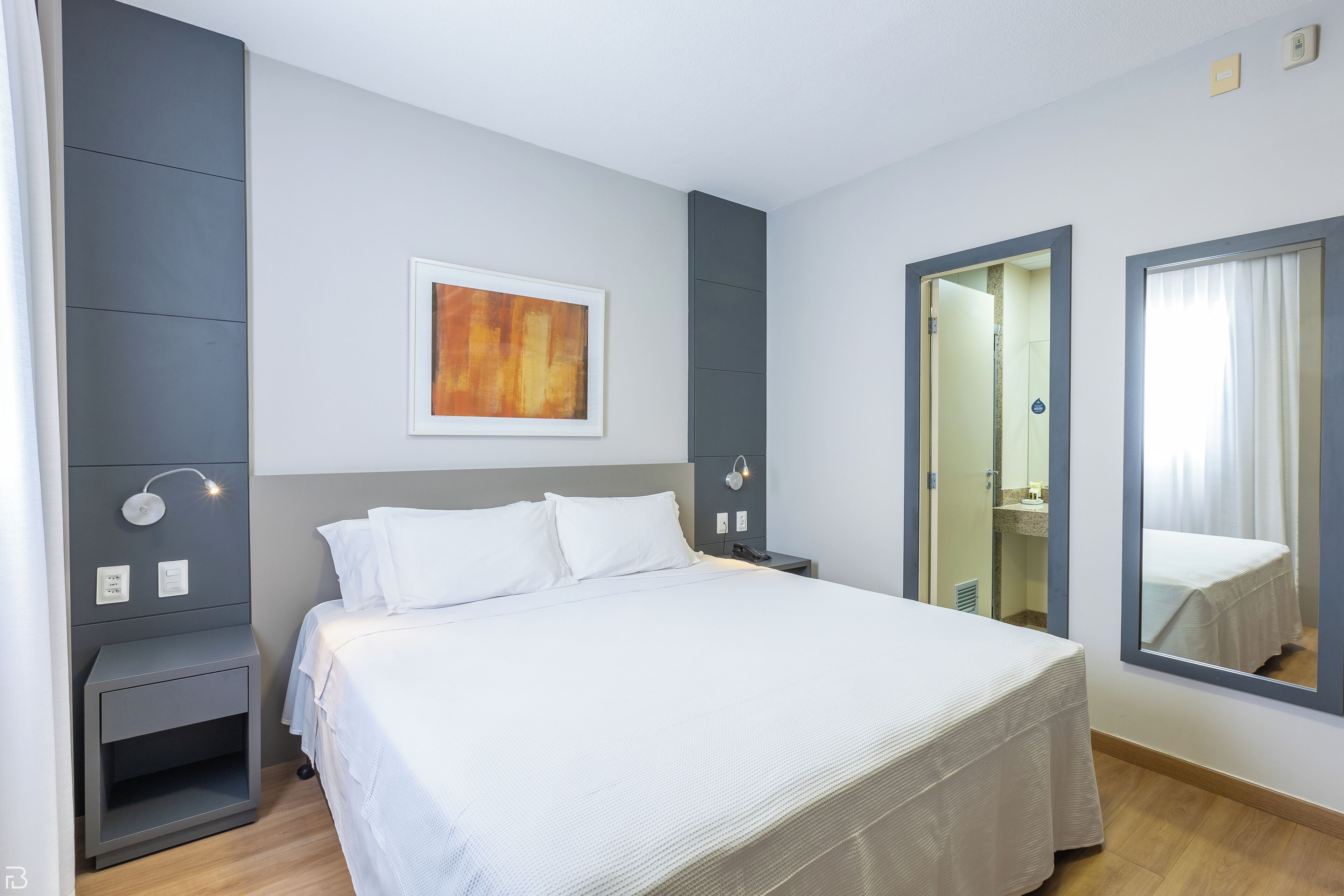Een moderne hotelkamer met een groot bed, nachtkastjes, een wandlamp, een spiegel en een kunstwerk in een lijst.