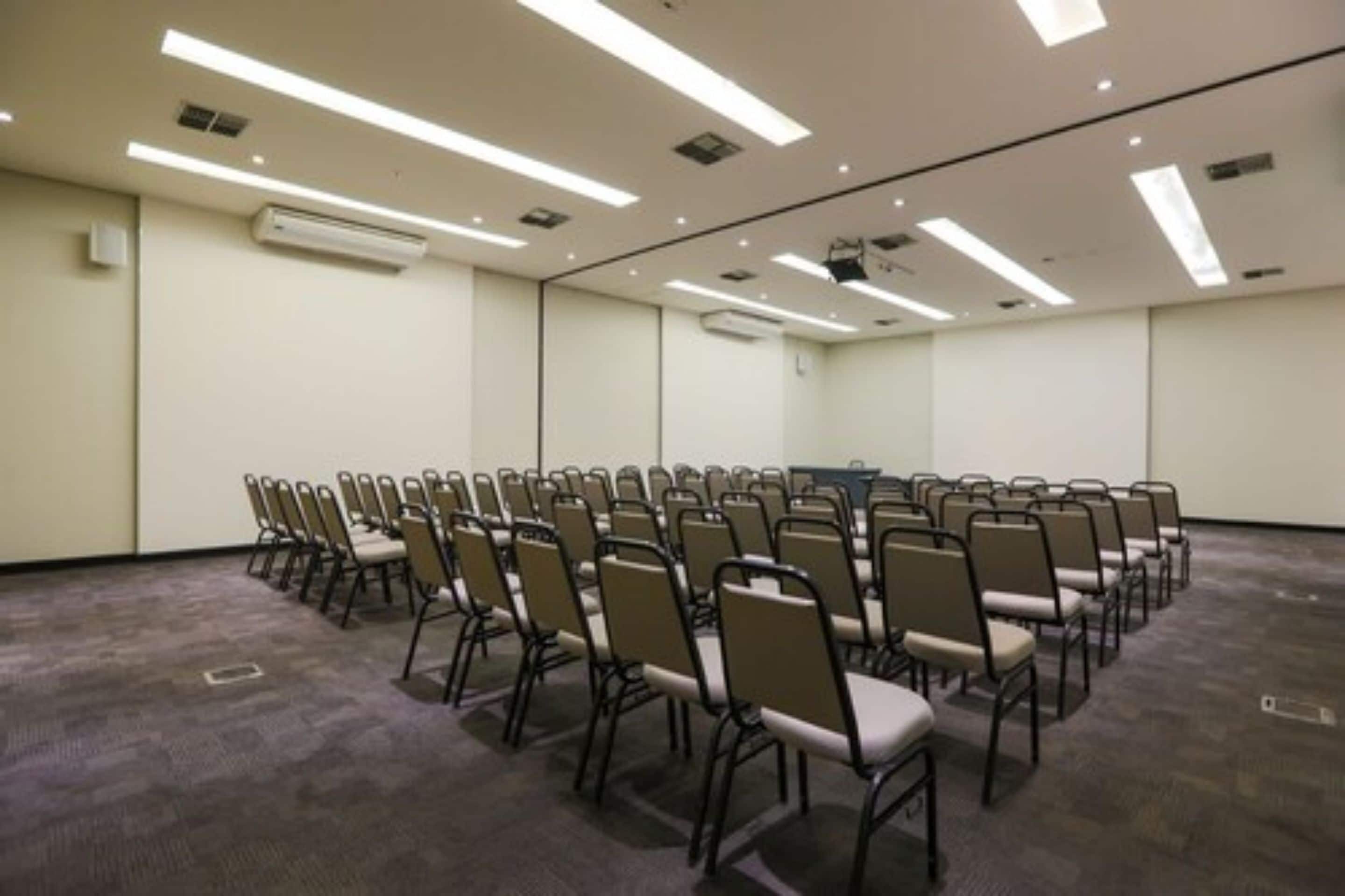 Un salón de conferencias con filas de sillas, luces en el techo y unidades de aire acondicionado.