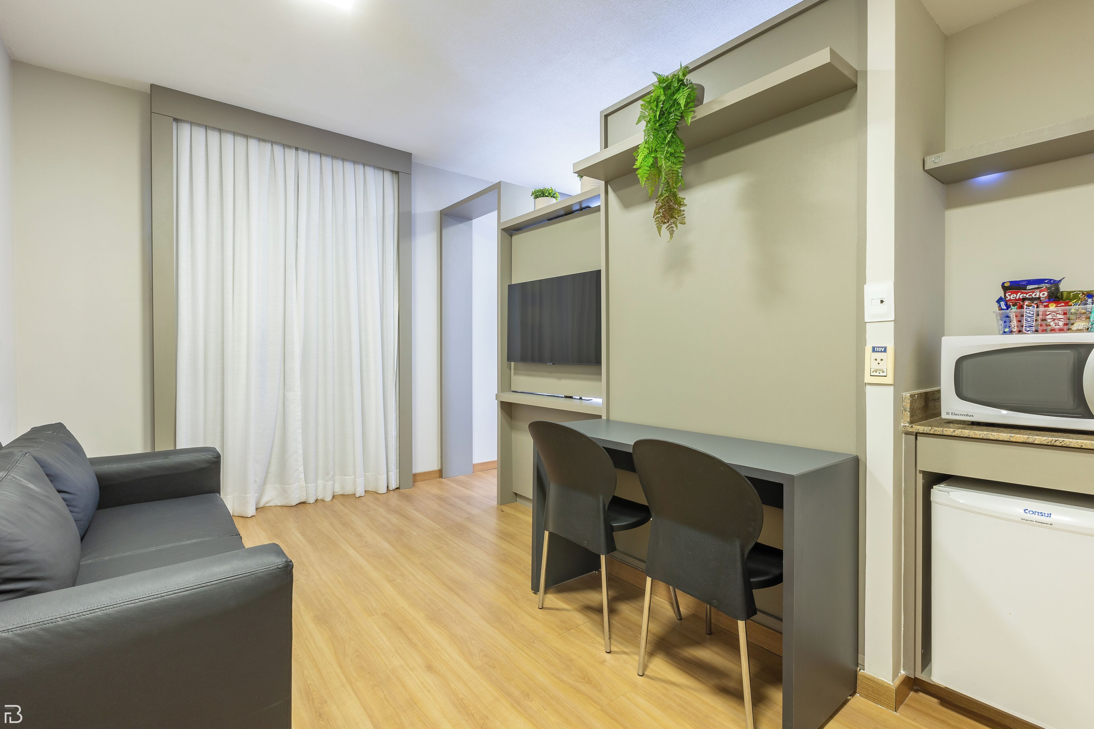 Una sala de estar moderna con un sofá gris, un escritorio con dos sillas, un televisor de pantalla plana, un refrigerador pequeño y una planta en maceta.
