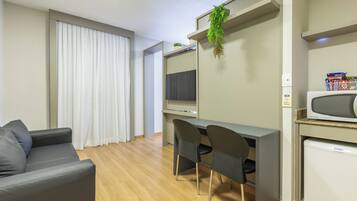Una sala de estar moderna con un sofá gris, un escritorio con dos sillas, un televisor de pantalla plana, un refrigerador pequeño y una planta en maceta.