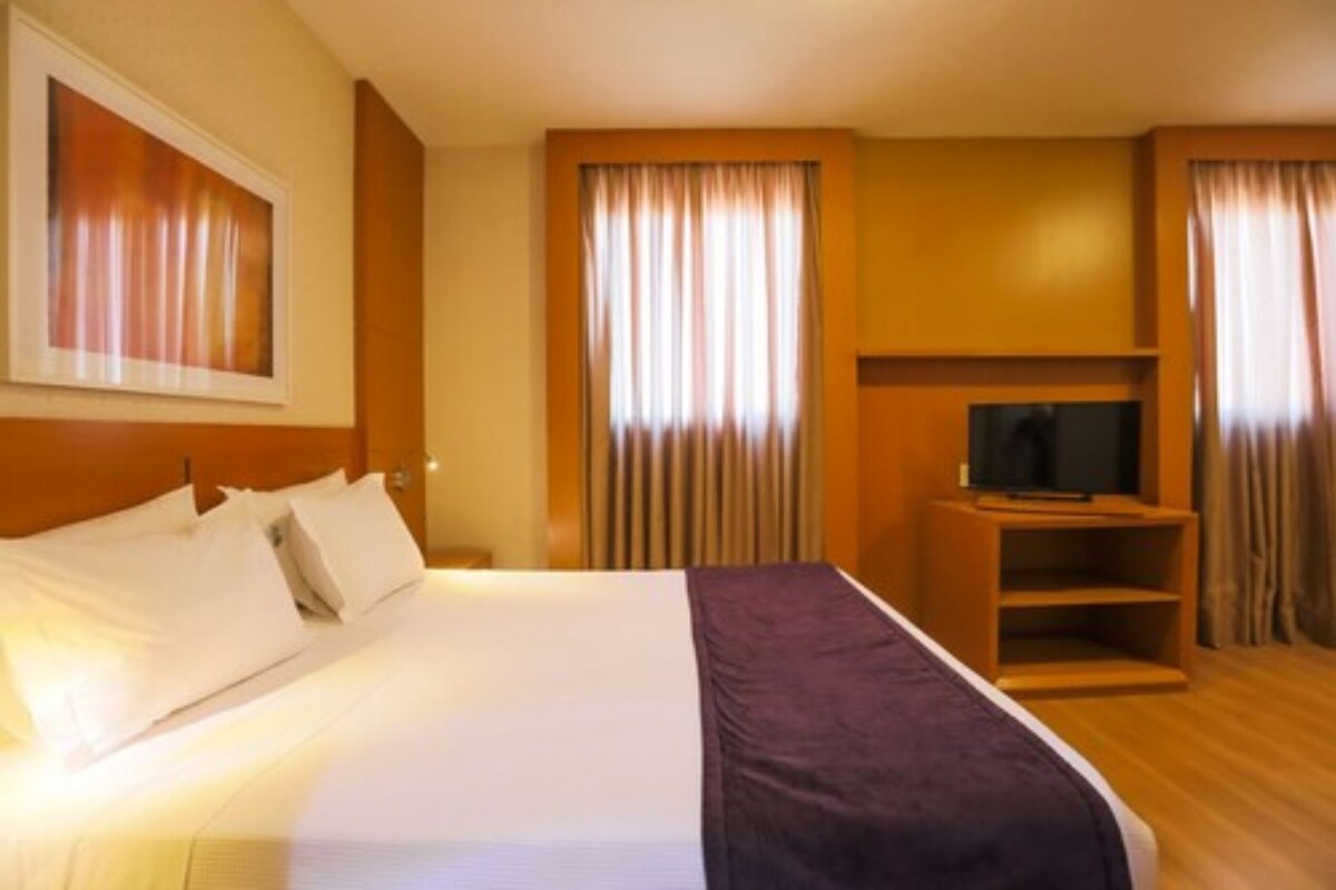 Una habitación de hotel con una cama, un televisor sobre un soporte de madera y cortinas en las ventanas.