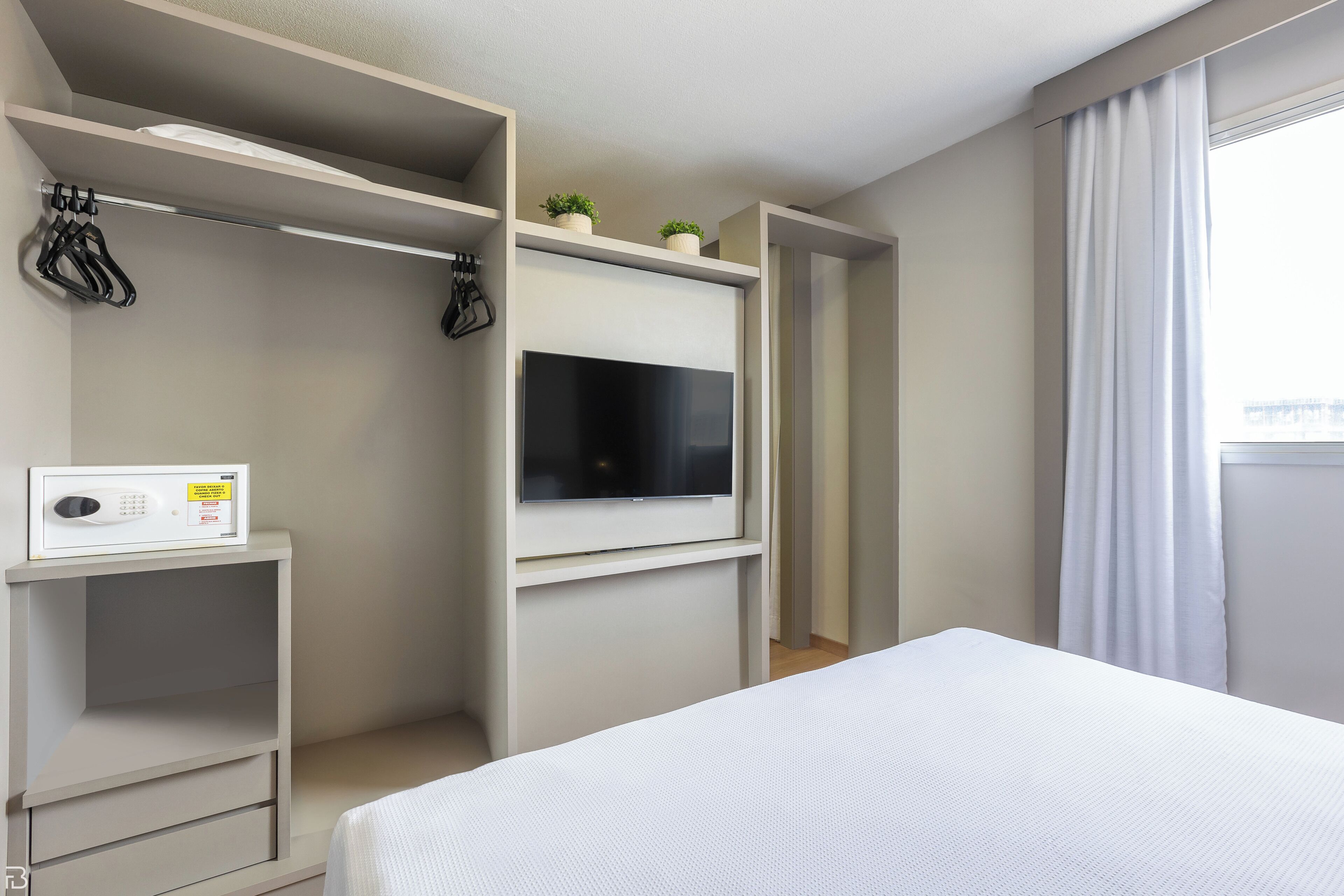 Habitación de hotel con cama, televisión montada en la pared, una estantería pequeña con microondas y ventana con cortinas.