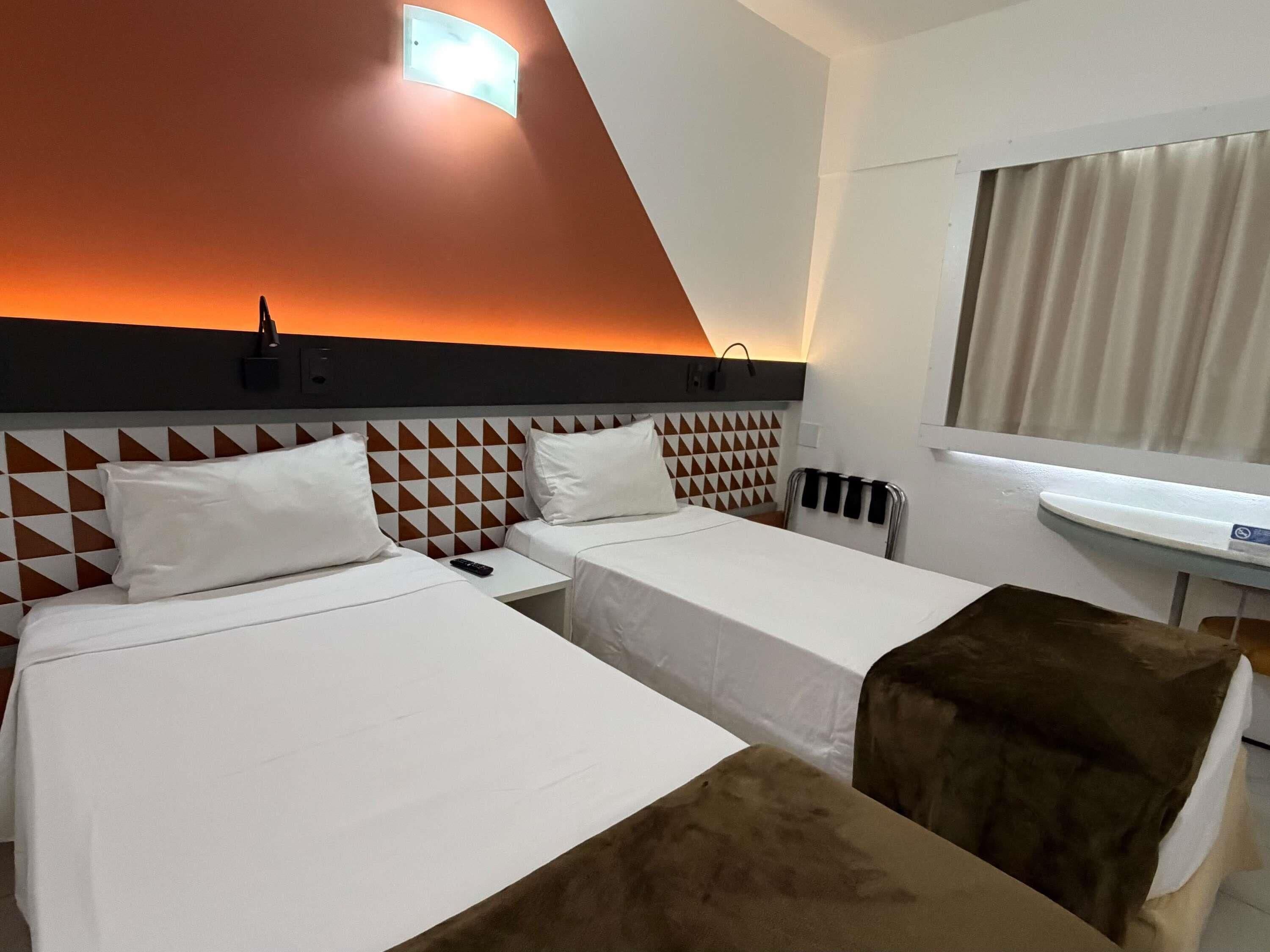 A hotel room with two beds, a headboard with a geometric pattern, a bedside table, a nightstand, a wall-mounted light, a window with curtains, and a small sink.