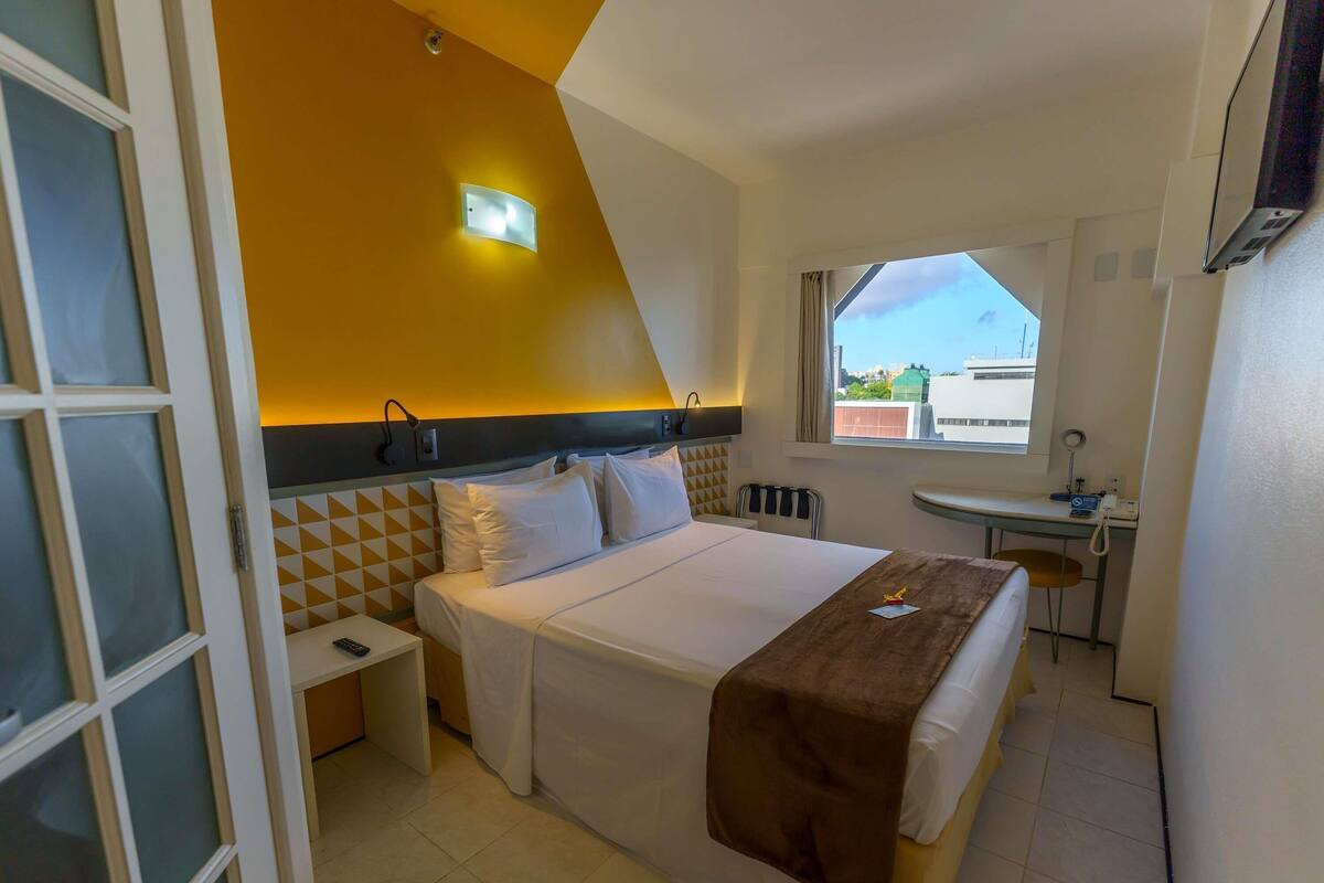 A hotel room with a bed, bedside table, lamp, window, and a view of the city.