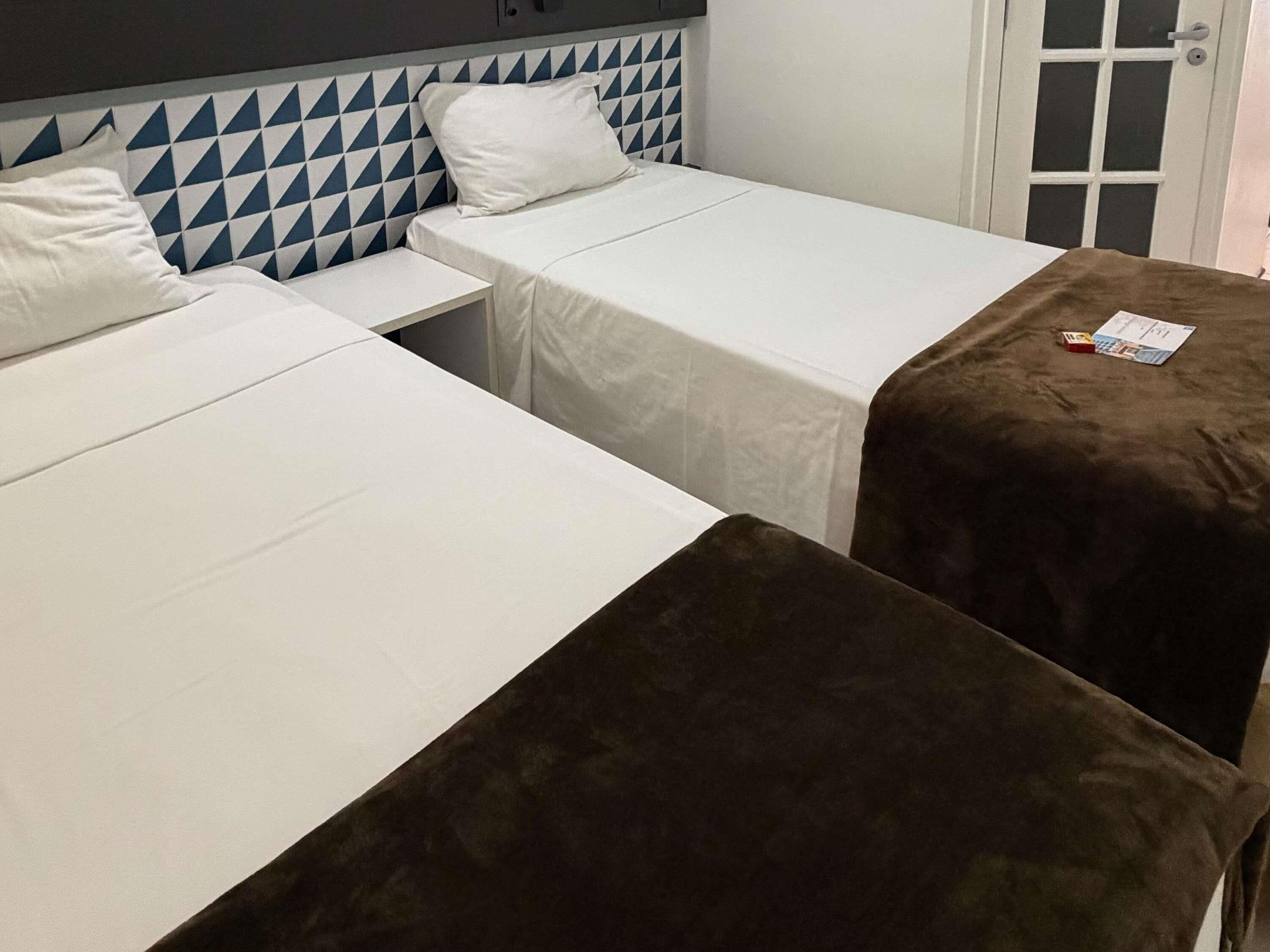 Quarto com duas camas, uma com colcha branca e a outra com colcha marrom. Há uma porta com painel de vidro e moldura branca.