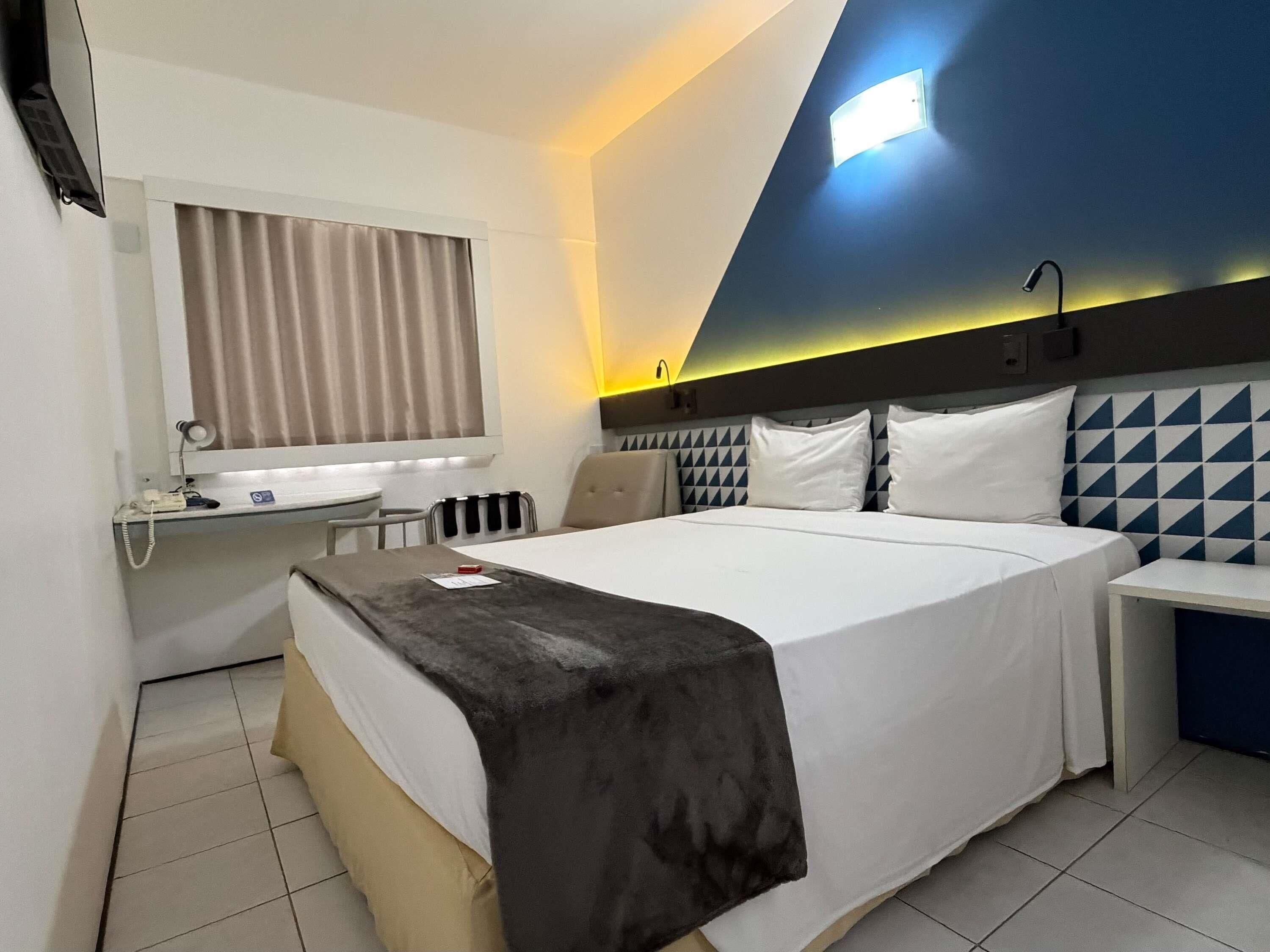 Una habitación de hotel con cama, mesita de noche, escritorio con silla, espejo y ventana con cortinas.