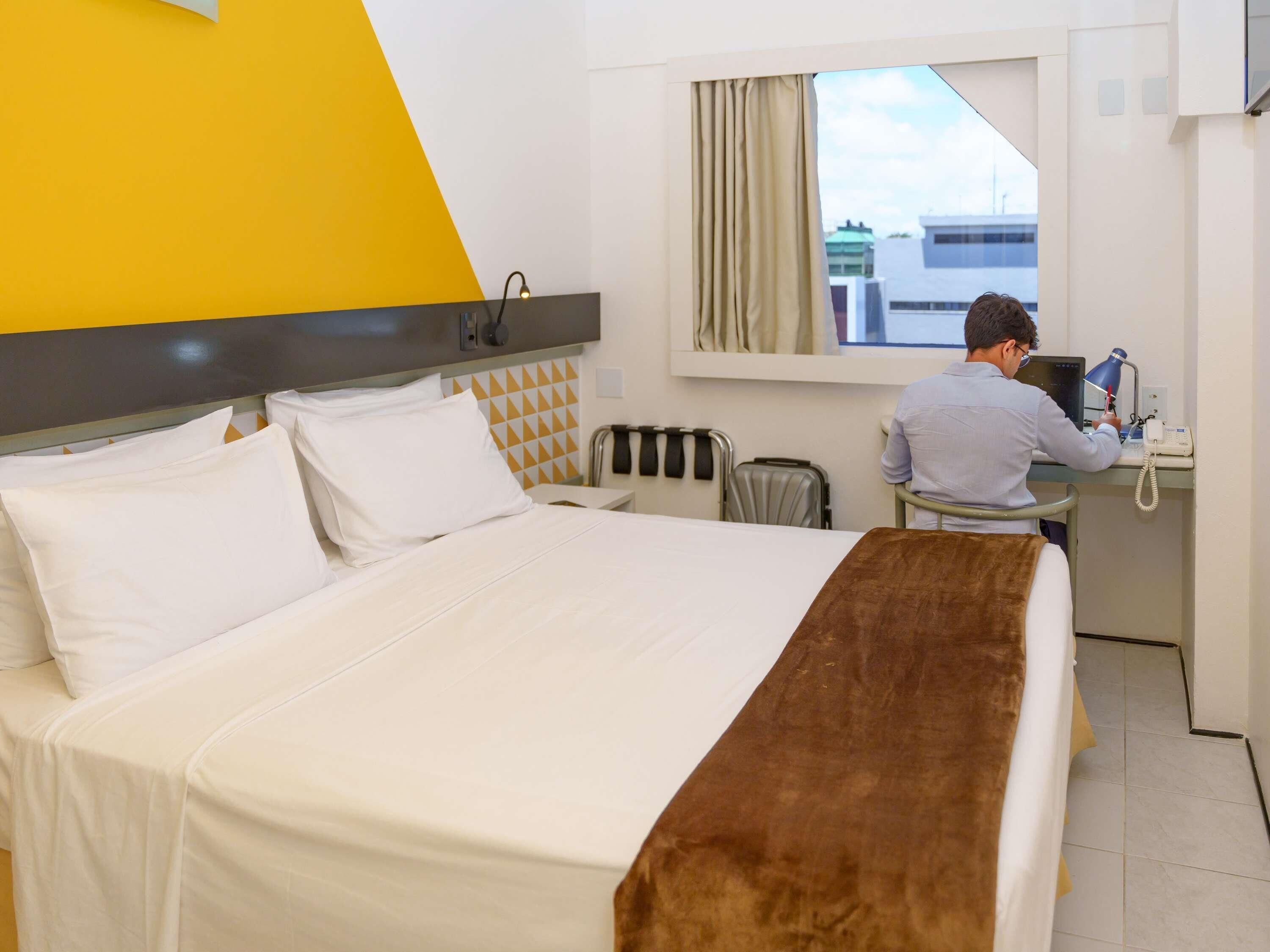 Ein Hotelzimmer mit einem Bett, einem Schreibtisch, einem Stuhl, einem Fenster mit Vorhängen und Blick auf Gebäude.