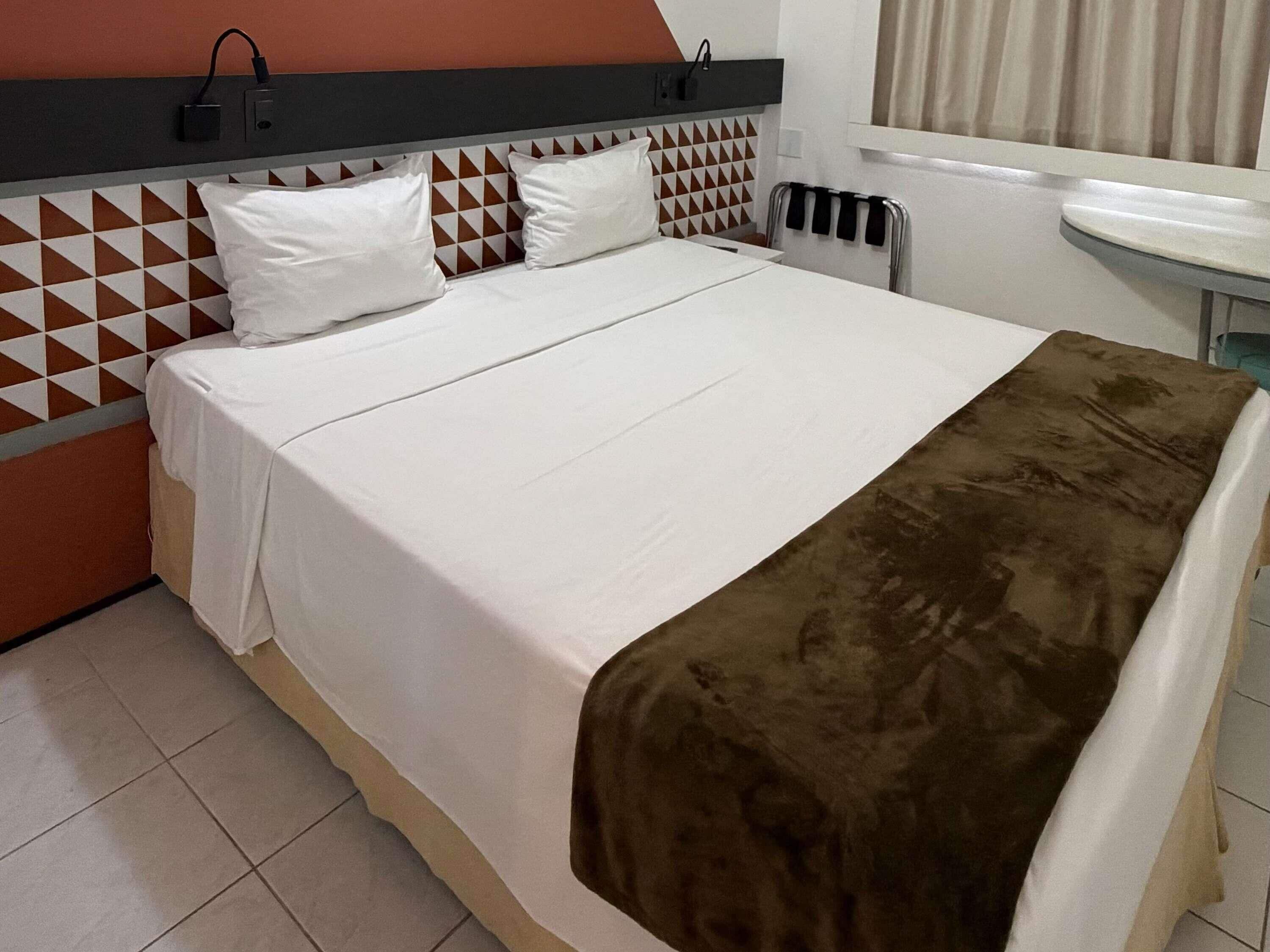 Habitación de hotel con cama, mesita de noche, radiador y ventana con cortinas.