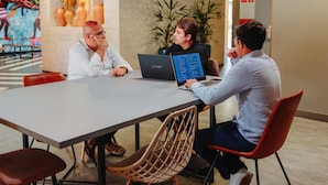 Ein Konferenzraum mit Tisch, Stühlen und drei Personen, die Laptops benutzen.