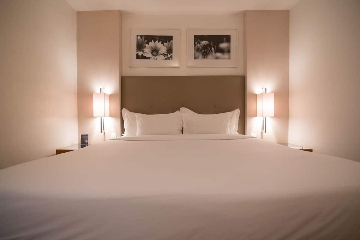 A large bed with white linens, two wall-mounted lamps, and two framed floral photographs above the bed.
