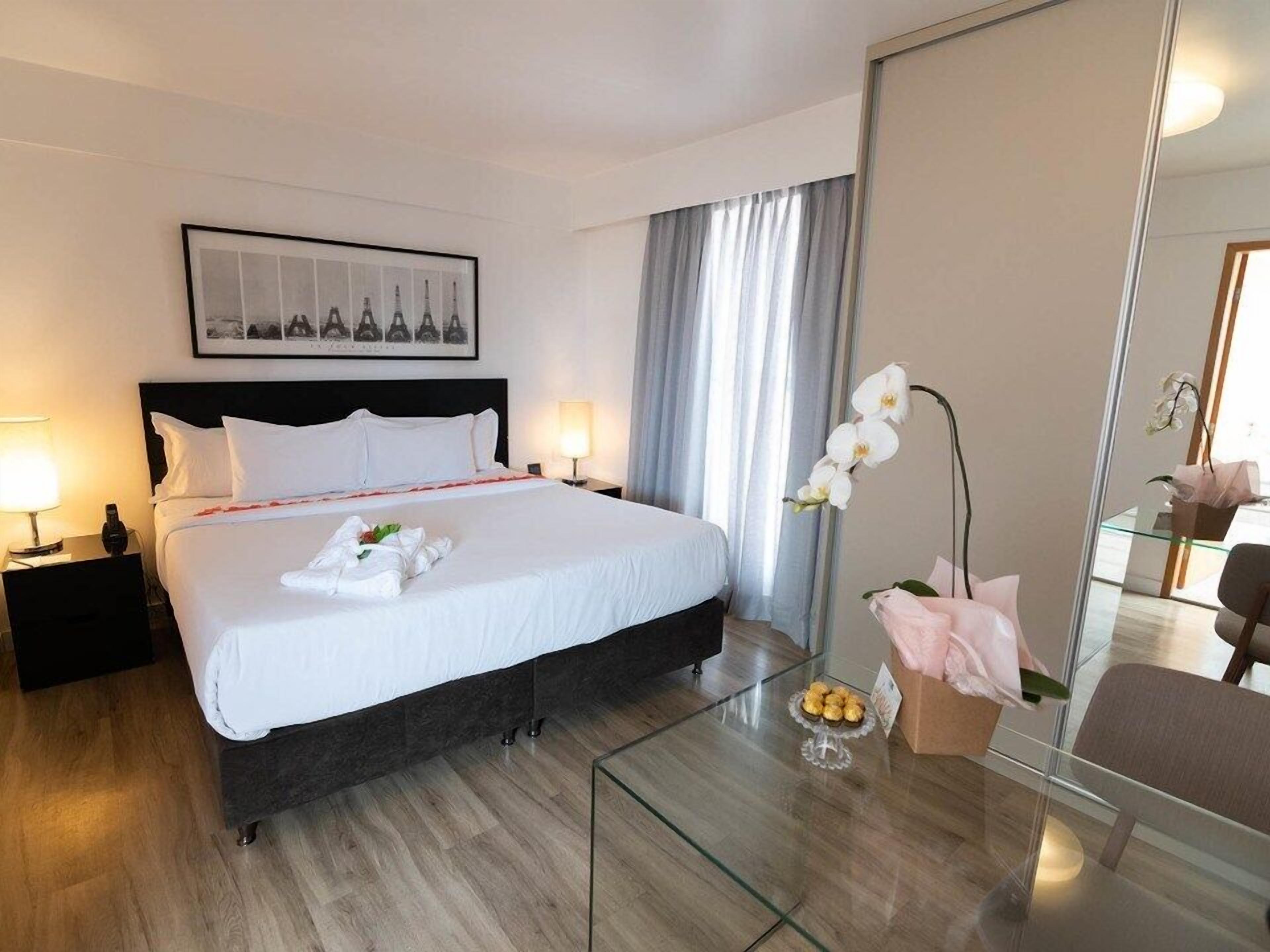 Una habitación de hotel con cama, mesita de noche, mesa de centro de vidrio, espejo y un cuadro en la pared.