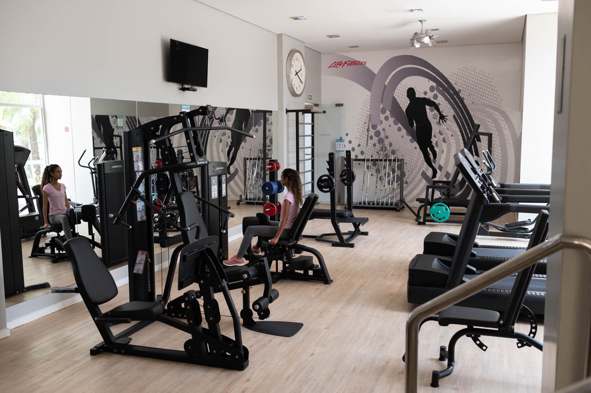 Uma academia com diversos equipamentos de exercício, um relógio e uma parede com um mural sobre fitness.