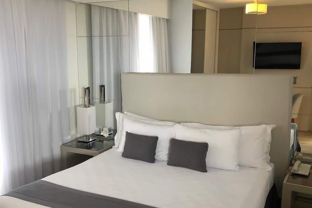 A modern hotel room with a large bed, a bedside table with a lamp, a flat-screen TV mounted on the wall, and a large window with curtains.