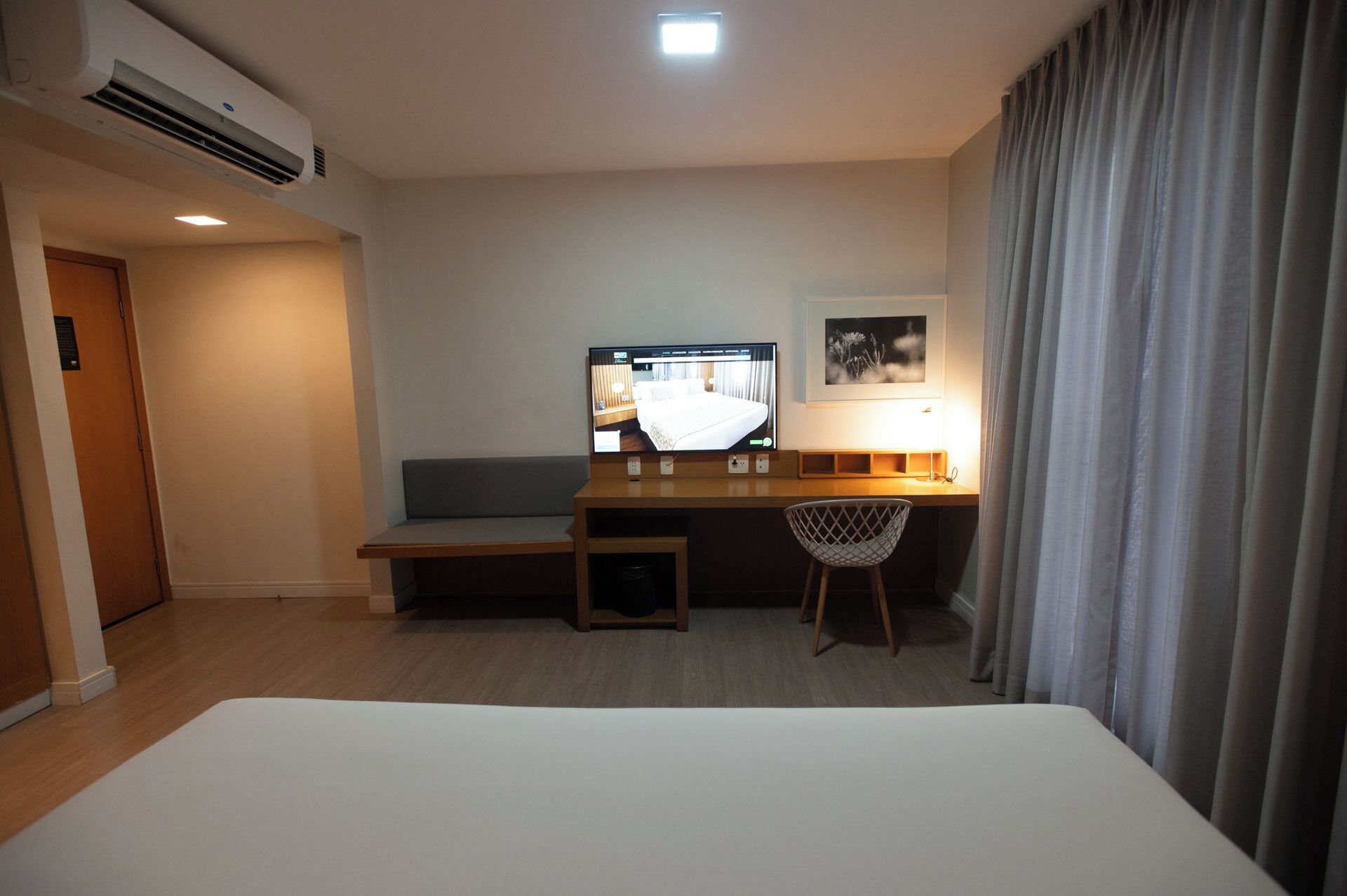 Habitación de hotel con cama, escritorio con computadora, una silla, televisor y ventana con cortinas.