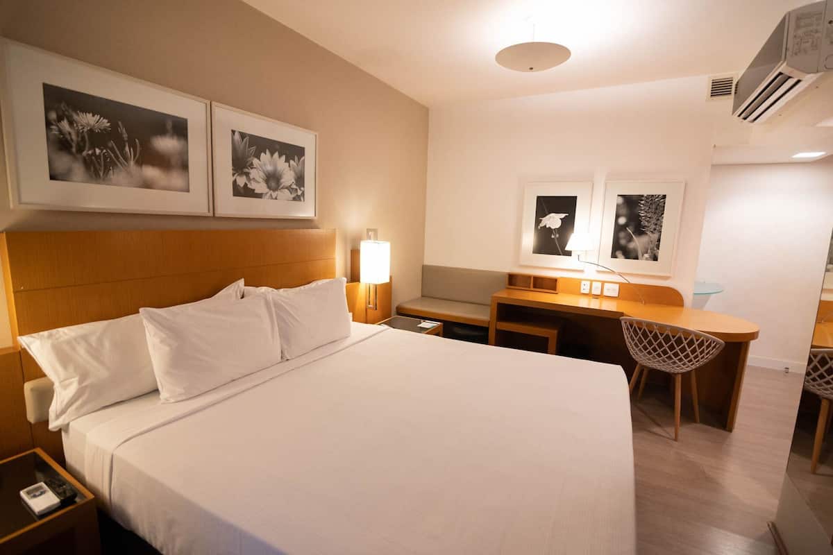 A hotel room with a bed, bedside table, desk, chair, and wall-mounted artwork.