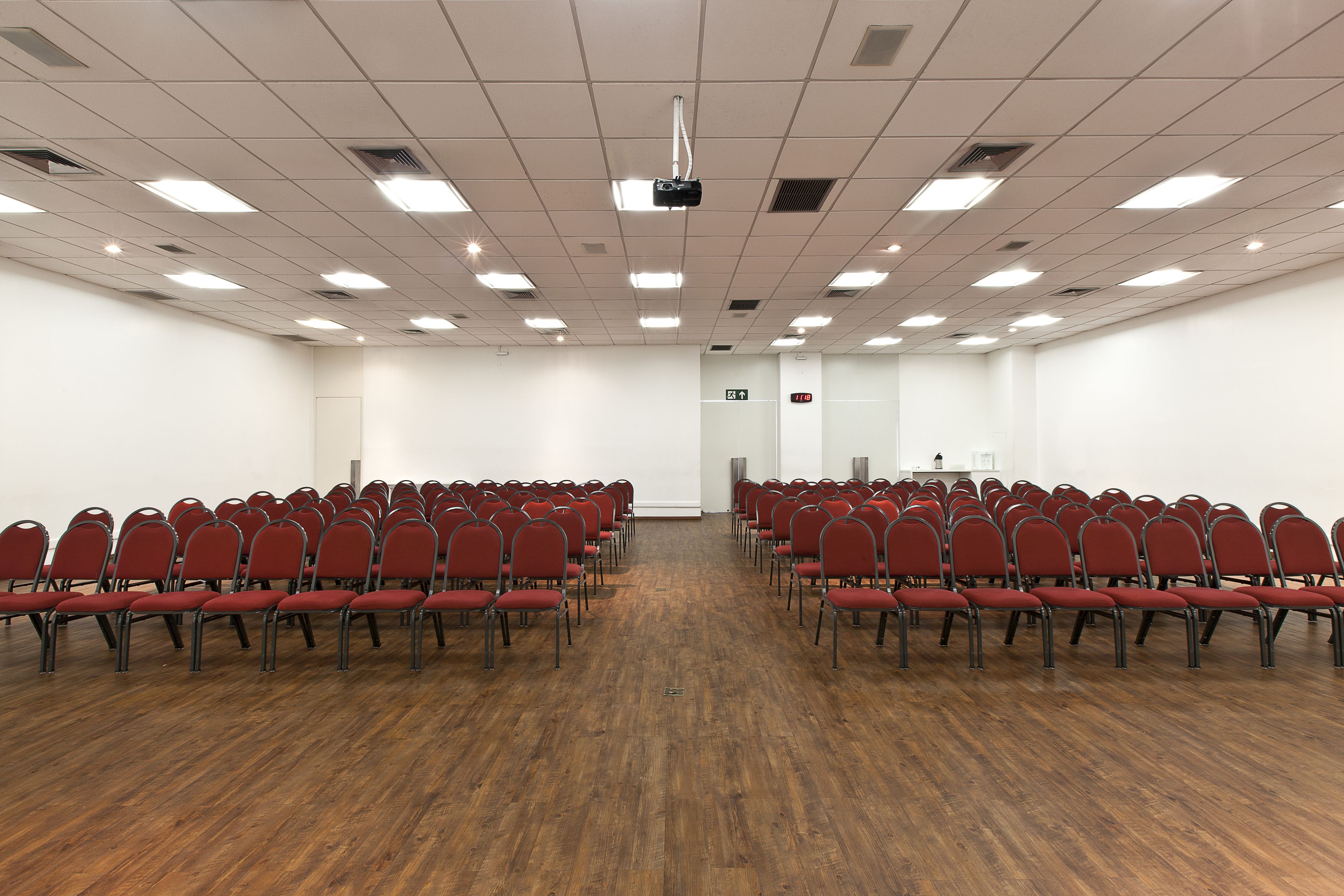 Un salón de conferencias con filas de sillas rojas, un proyector en el techo y suelo de madera.