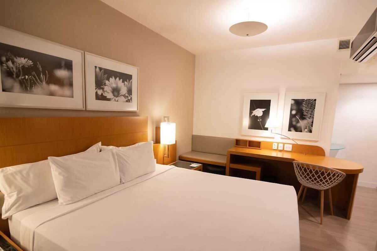 A hotel room with a bed, bedside table, lamp, desk, chair, and two framed pictures on the wall.