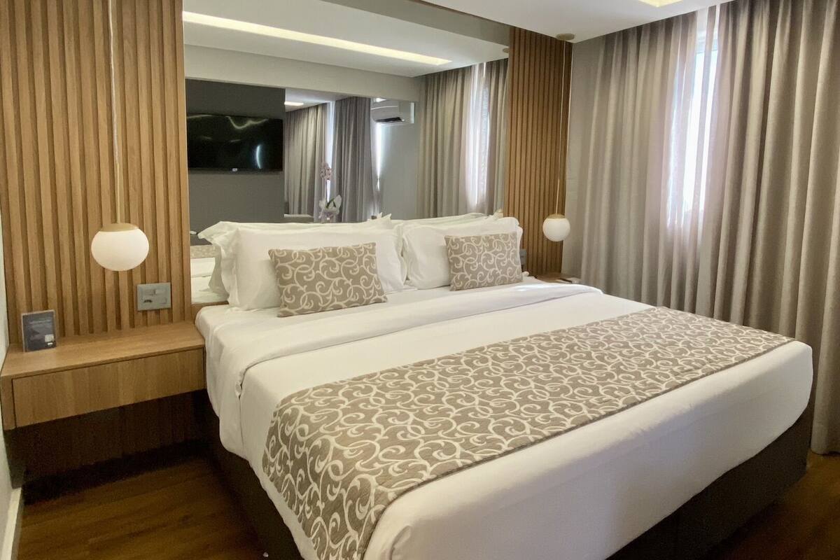 A modern hotel room with a large bed, wooden paneling, a flat-screen TV, and a large window with curtains.