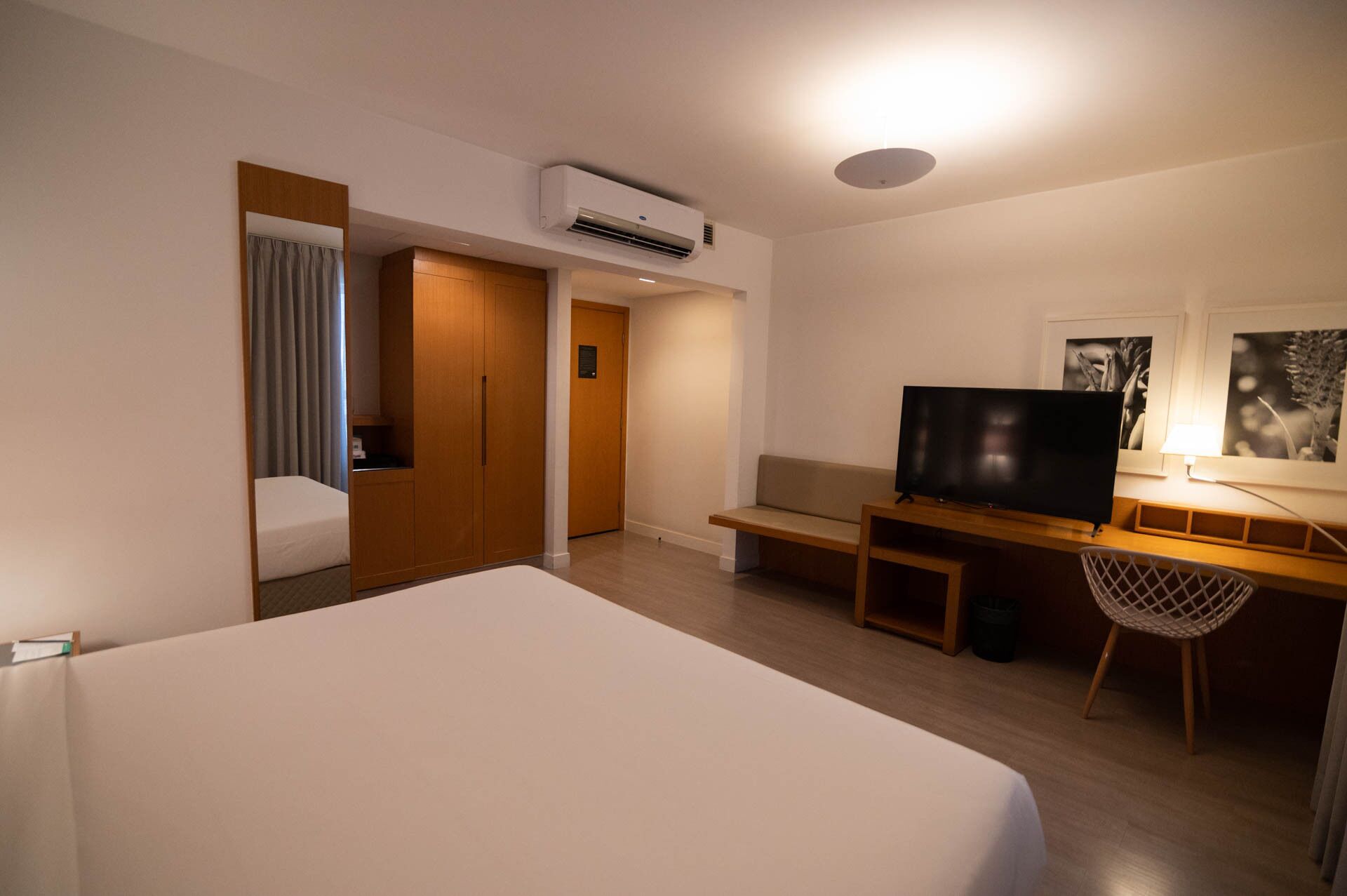 A hotel room with a large bed, a desk with a TV, a chair, and an air conditioning unit.