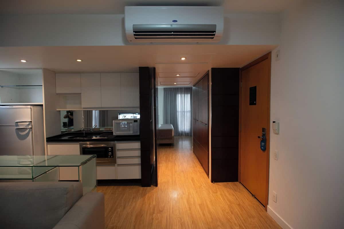 A modern kitchen with a refrigerator, microwave, and oven. There is a glass table and a sofa in the living area. The room has wooden flooring and a door leading to another room.