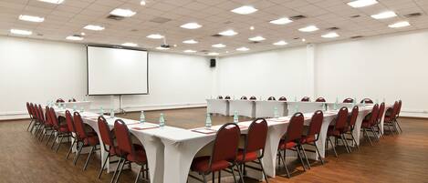 Un salón de conferencias con largas mesas, sillas rojas, manteles blancos y una pantalla de proyector.