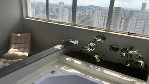 Een moderne badkamer met een groot bad, een comfortabele stoel en uitzicht op een stadslandschap door grote ramen.