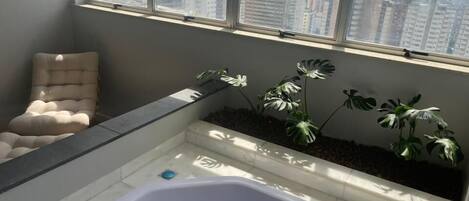A modern bathroom with a large bathtub, a cushioned chair, and a view of a cityscape through large windows.