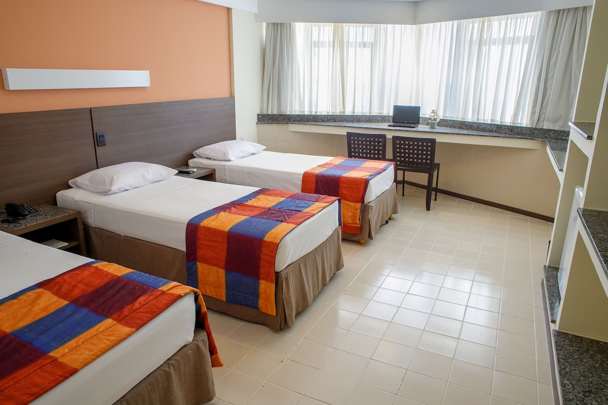 A hotel room with three beds, a desk with a laptop, and a window with curtains.